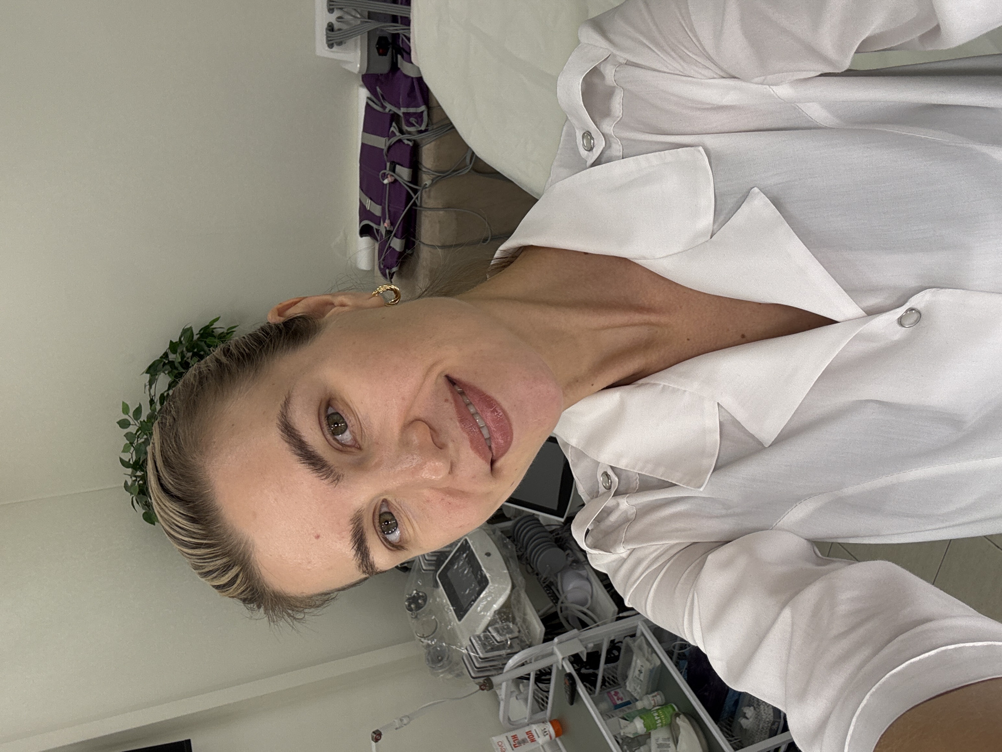 A person is captured in a selfie, with the image rotated 90 degrees counter-clockwise. The subject is a woman with light-colored hair, styled away from her face, revealing highlights. She has prominent eyebrows, brown eyes, and a slight, friendly smile showing her teeth. She wears a white collared shirt, possibly a uniform or scrub top, and a small gold hoop earring in her visible ear.
The background suggests an indoor, clinical, or medical setting. To her left, a plain white wall is visible. Behind her and to her right, various medical equipment and supplies are arranged. This includes a machine with a digital screen and multiple tubes or wires attached, along with a white plastic caddy containing several bottles and containers. Purple fabric with straps, possibly part of medical equipment or a patient gown, hangs in the upper background. A small, leafy green plant, possibly artificial, is positioned near her head.
The activity appears to be a self-portrait taken in what looks like a professional healthcare environment. Her expression is pleasant and composed.
Visible text on bottles in the background includes "ГИДРОПЕРИТ" (GIDROPERIT, likely hydrogen peroxide) and "ЭВИТОЛ" (EVITOL). Other labels are present but illegible due to distance and resolution. No specific time of day or weather cues are discernable.