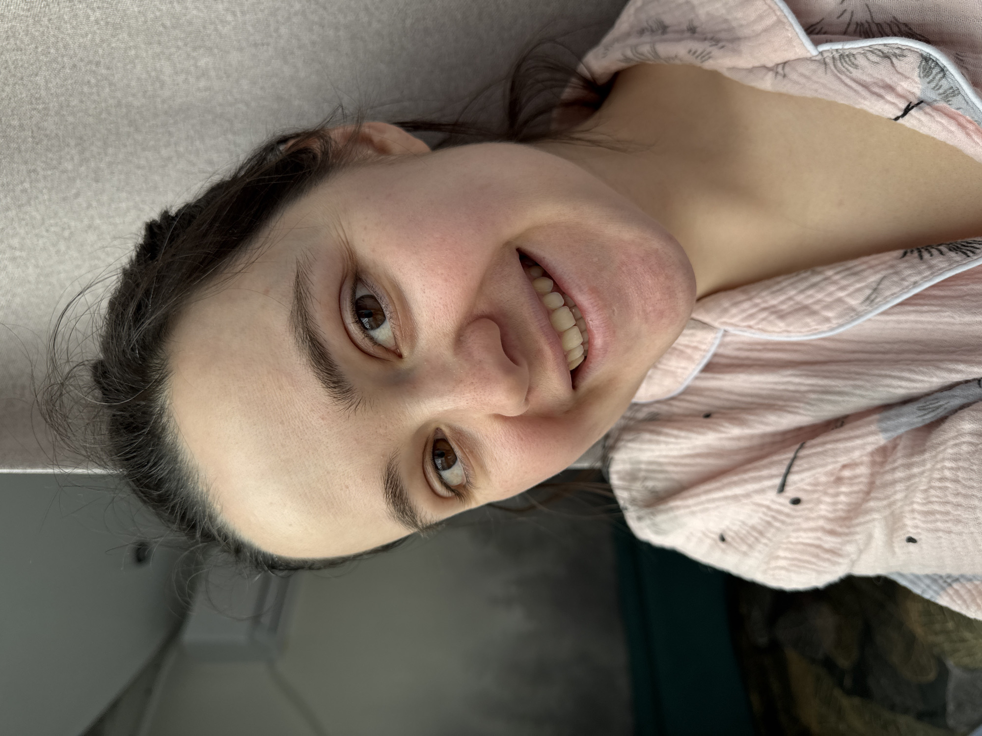 A woman is pictured from the chest up in a close-up shot. She has brown hair and eyes and is wearing a pink and white patterned pajama top. She is smiling and looking at the camera. The background is a mix of gray and dark green. It is unclear if the location is in Izhevsk, Russia.