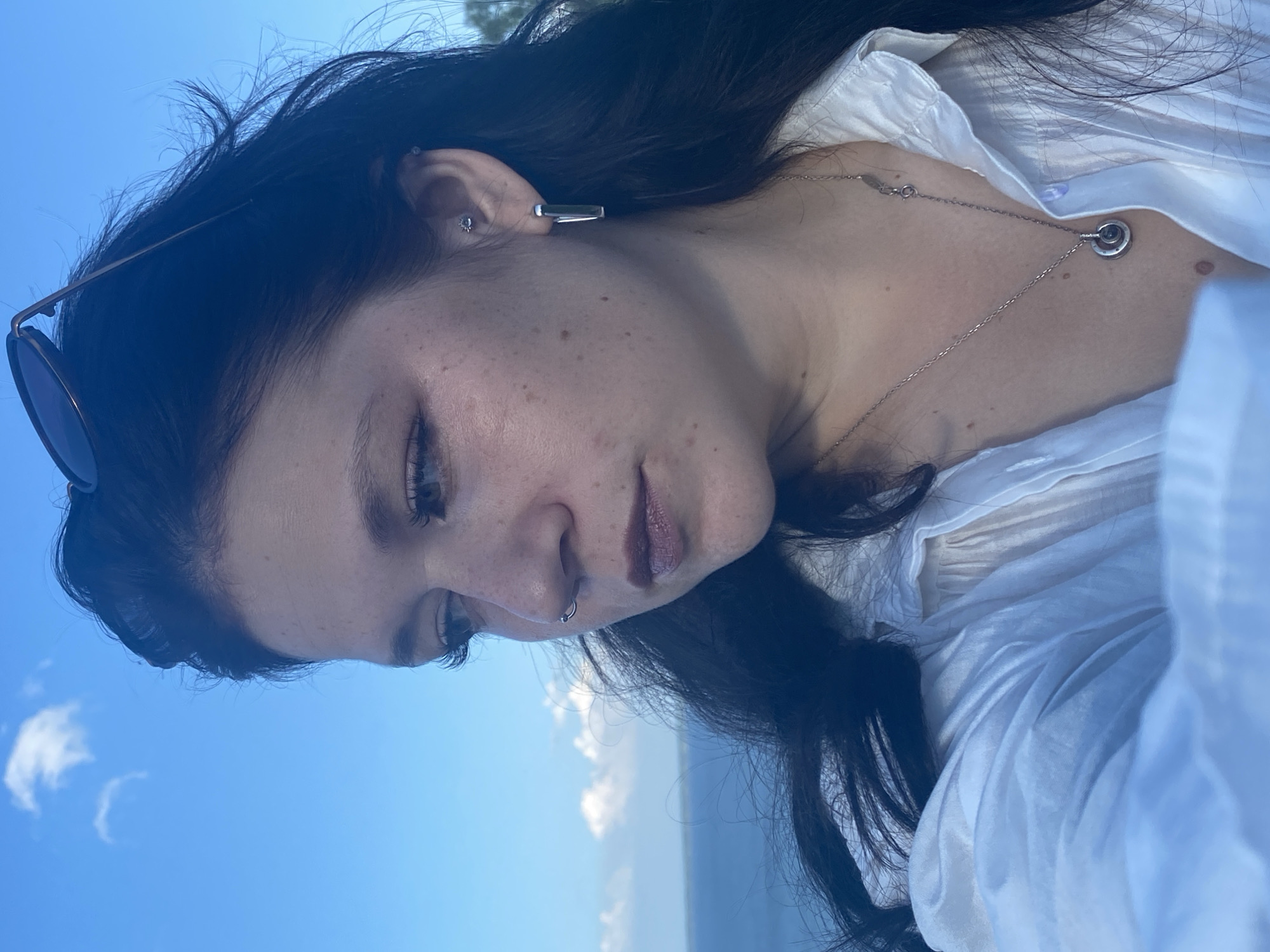 A close-up, profile shot of a young woman with dark hair is captured against a bright blue sky with scattered clouds. She is wearing a white collared shirt, a silver necklace with a circular pendant, and a silver hoop earring in her left ear. She also has a silver ring piercing in her right nostril and a small stud piercing in her left ear. Sunglasses are perched on top of her head. The background shows a body of water and a distant shoreline, suggesting an outdoor setting possibly near the coast. The image does not provide any information to determine a specific location within Kostovaty, Russia.