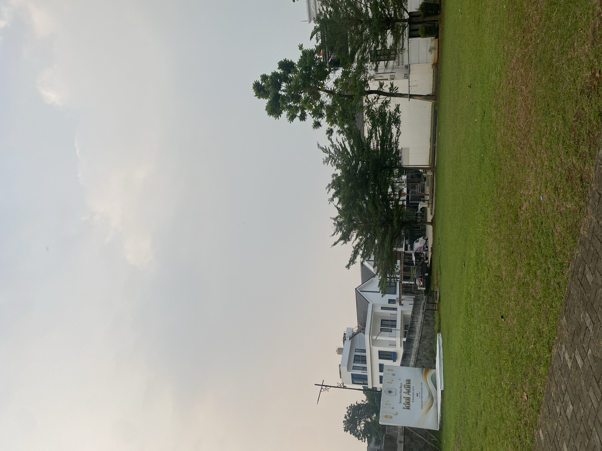 The image depicts a street scene in Cibinong, Indonesia, near the Sentul City area, characterized by modern white houses with blue accents and a large expanse of manicured green lawn in the foreground. A large banner that reads "Selamat Hari Raya Idul Adha" (Happy Eid al-Adha) is prominently displayed on the left side of the image, accompanied by decorative elements resembling crescent moons and stars, and the date "10 Dzulhijjah 1443 H". Several trees, some tall and slender, others with fuller canopies, are interspersed between the houses and along the property lines. A street light stands to the left of the houses, and a paved walkway is visible at the bottom of the frame. The sky is overcast, suggesting a hazy or cloudy day. A few cars, including a white van and a dark-colored SUV, are parked in the background, near the houses. The overall impression is of a residential development or a community area, possibly decorated for a religious holiday.