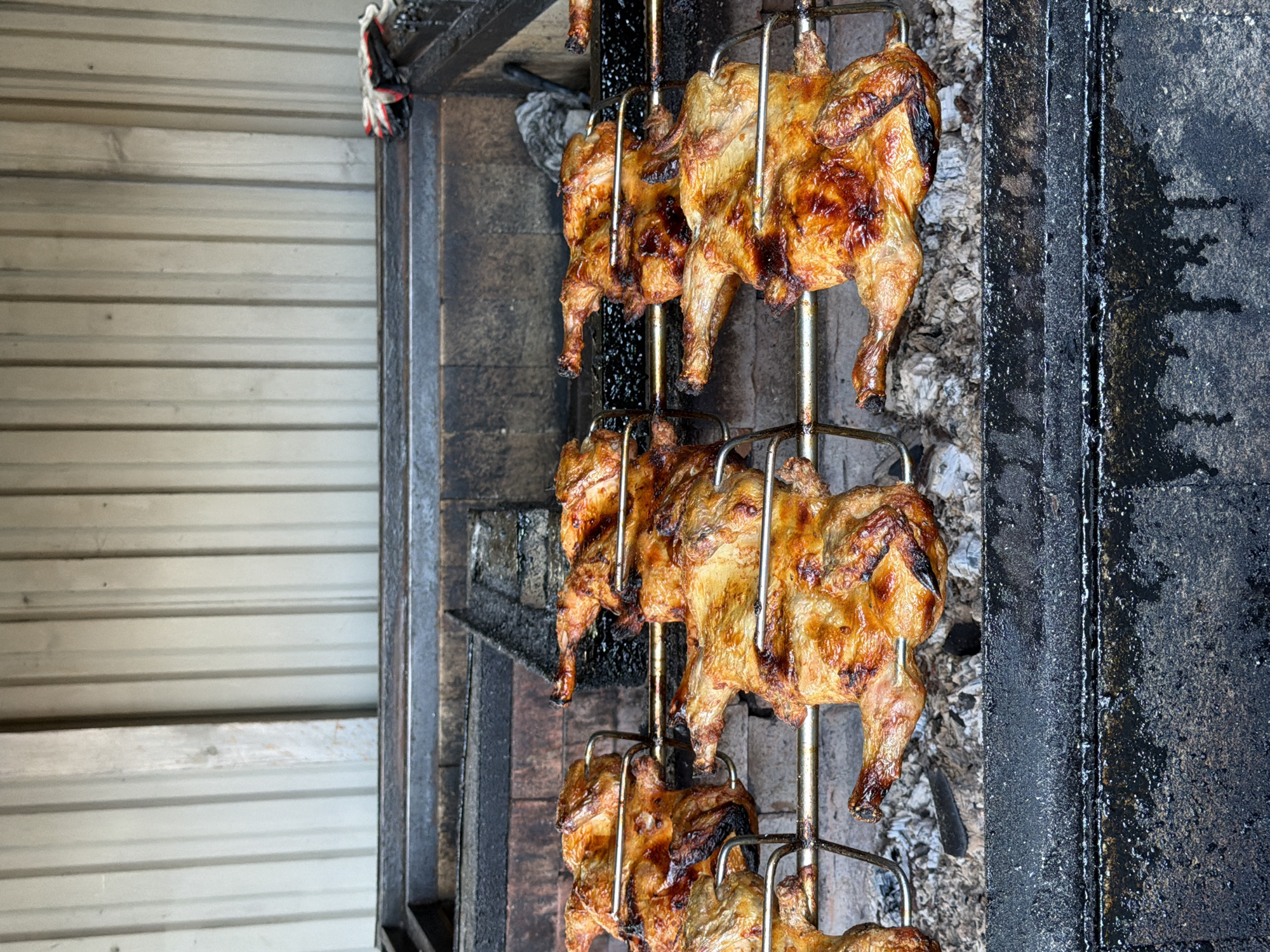 A close-up, low-angle shot captures several whole chickens rotating on a spit, likely at an outdoor food stall or restaurant in Sochi, Russia. The chickens are golden brown and appear to be well-seasoned, with some areas showing a darker, crispy char. They are skewered on metal rods, suspended above what appears to be a grill or fire pit, with visible embers and ashes beneath the bottom row of chickens. To the left of the spit, a corrugated metal wall provides a textured background. To the right, a dark, possibly cast-iron surface, stained with grease and residue, suggests the cooking apparatus. The lighting is natural, implying it is daytime. There is no visible text in the image. The overall impression is one of delicious, freshly roasted chicken being prepared for customers.