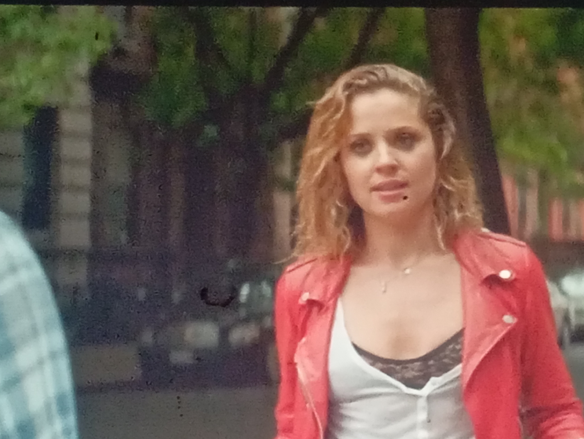 The image shows a medium shot of a light-skinned woman with blonde, wavy hair. She is wearing a red leather jacket over a white top with a black lace camisole underneath. A small, silver necklace is visible on her chest. The woman has a mole on her cheek. She is looking towards the left of the frame. In the background, trees and buildings suggest an outdoor urban setting, possibly a street in Sofiivska Borshchahivka, Ukraine, though this cannot be definitively confirmed from the image alone. To the left of the woman, a portion of another person's shoulder and arm, wearing a blue and white plaid shirt, is visible, indicating a possible interaction or shared space. The overall image has a slightly grainy texture.