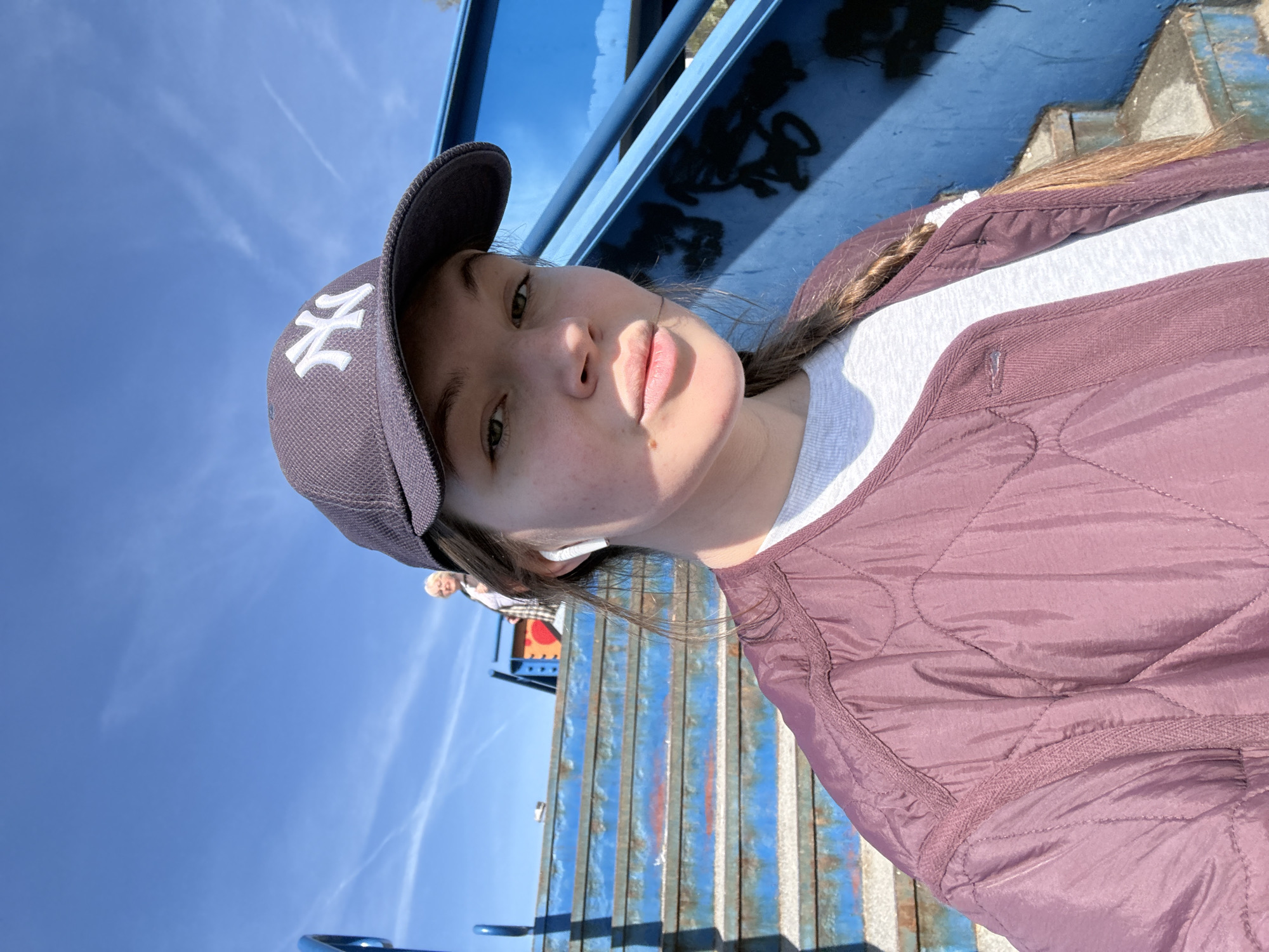 The image shows a young woman wearing a dark baseball cap with the New York Yankees logo, a white t-shirt, and a quilted maroon jacket. She has fair skin, light brown hair pulled back in a braid, and is wearing an earbud in her left ear. She is outdoors, possibly at a playground or recreational area, as suggested by the blue metal structure and bars in the background. The sky is visible and appears blue. Based on the environment, the location could be in Vozdovac Urban Municipality, Serbia.