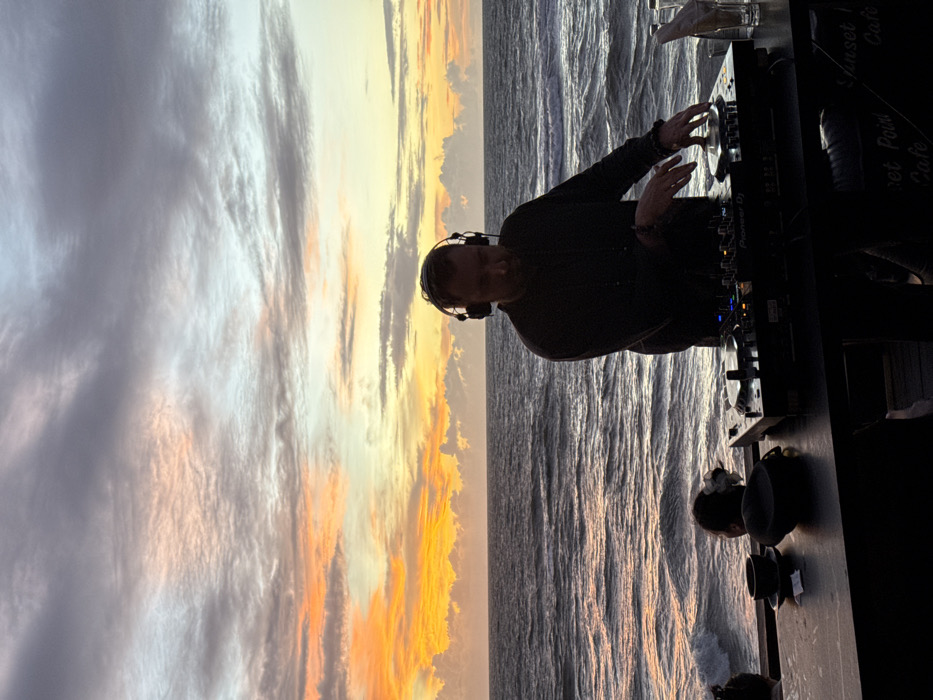 The image depicts an outdoor scene at a beachfront establishment, partially identified as "Sunset Point Cafe" by a sign in the lower right, located in Canggu, Indonesia, occurring during sunset. A male individual, centrally positioned, is operating a Pioneer DJ setup on a dark counter. He wears over-ear headphones and a dark long-sleeved garment, with both hands engaged on the mixer and players. To the left of the DJ equipment on the counter, a dark cup with a saucer and a dark hat are present. To the right, a glass bottle and a drinking glass containing a straw are visible. The background consists of the ocean with discernible waves extending to the horizon, beneath a sky characterized by strata of orange, yellow, pink, and grey clouds, consistent with a sunset. A portion of another individual's head, featuring a white flower, is visible in the lower-left corner below the counter.