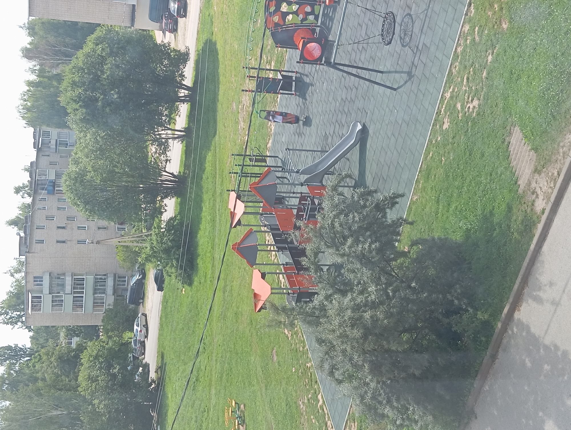The image depicts a playground in an urban environment, likely located in Suvorov, Russia, given the architectural style of the buildings and the general ambiance. In the background, there are multi-story apartment buildings with light-colored facades, characteristic of Soviet-era architecture. A line of trees separates the buildings from the foreground. The playground itself is a modern facility with a rubberized safety surface. It features various play structures including slides, swings, monkey bars, and climbing apparatus. A child is visible on a zipline, appearing to be in motion. The playground is bordered by green grass, and a paved area can be seen in the lower left of the image. The overall scene suggests a typical residential area with recreational facilities for children.
