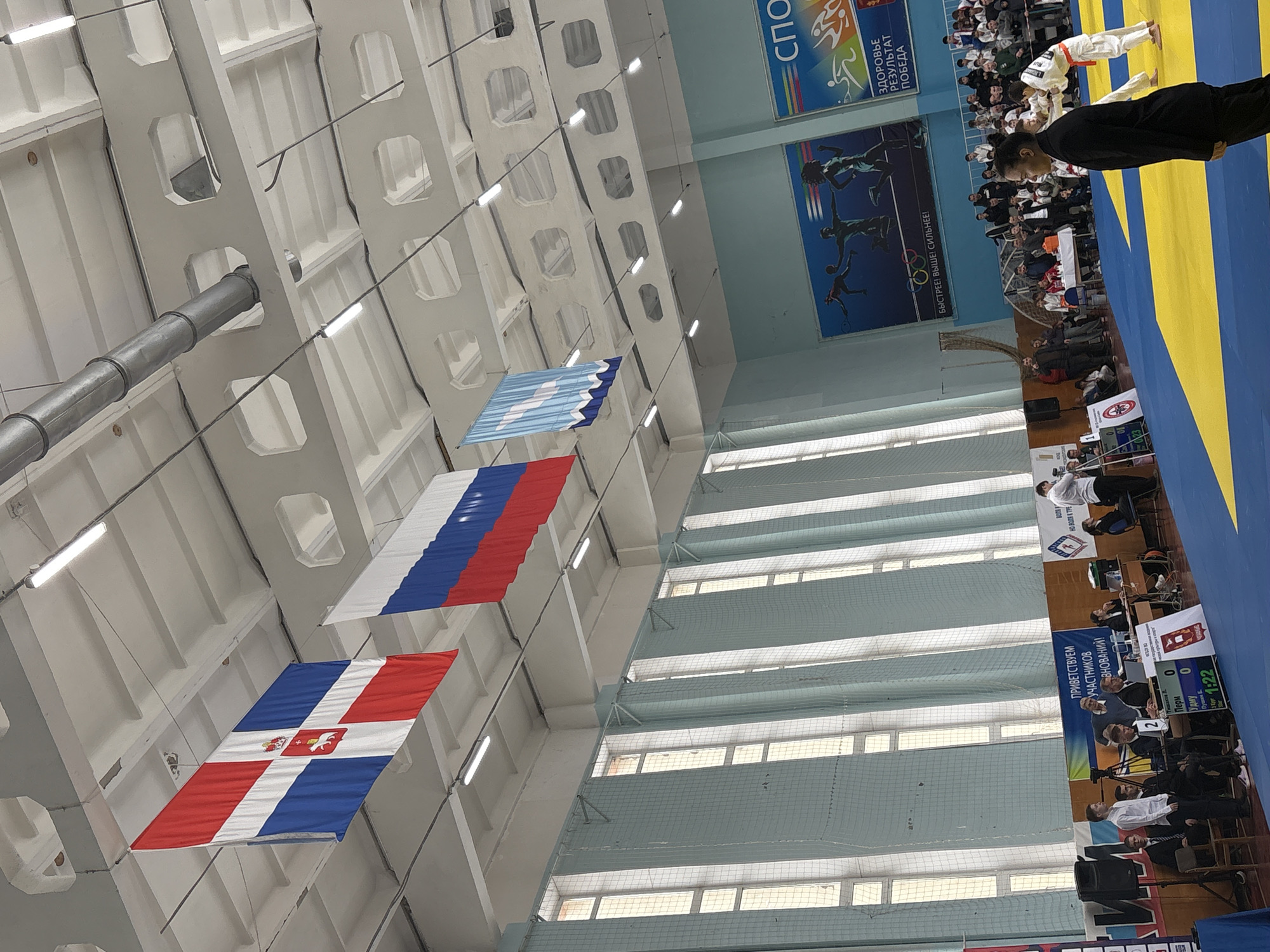 The image shows the interior of a sports hall in Chaikovsky, Russia, identifiable by the presence of the Perm region flag (red and white with a symbol in the center) and the Russian flag (white, blue, and red). The flags are hanging from the ceiling, which has a complex grid structure with lights. There are banners on the wall promoting sports and health. In the background, people are seated around a blue and yellow mat, where a martial arts competition appears to be taking place. A person in a black suit stands near the mat, possibly an official. At the right, there is a table with officials monitoring the contest. A scoreboard indicates the time and scores of participants.