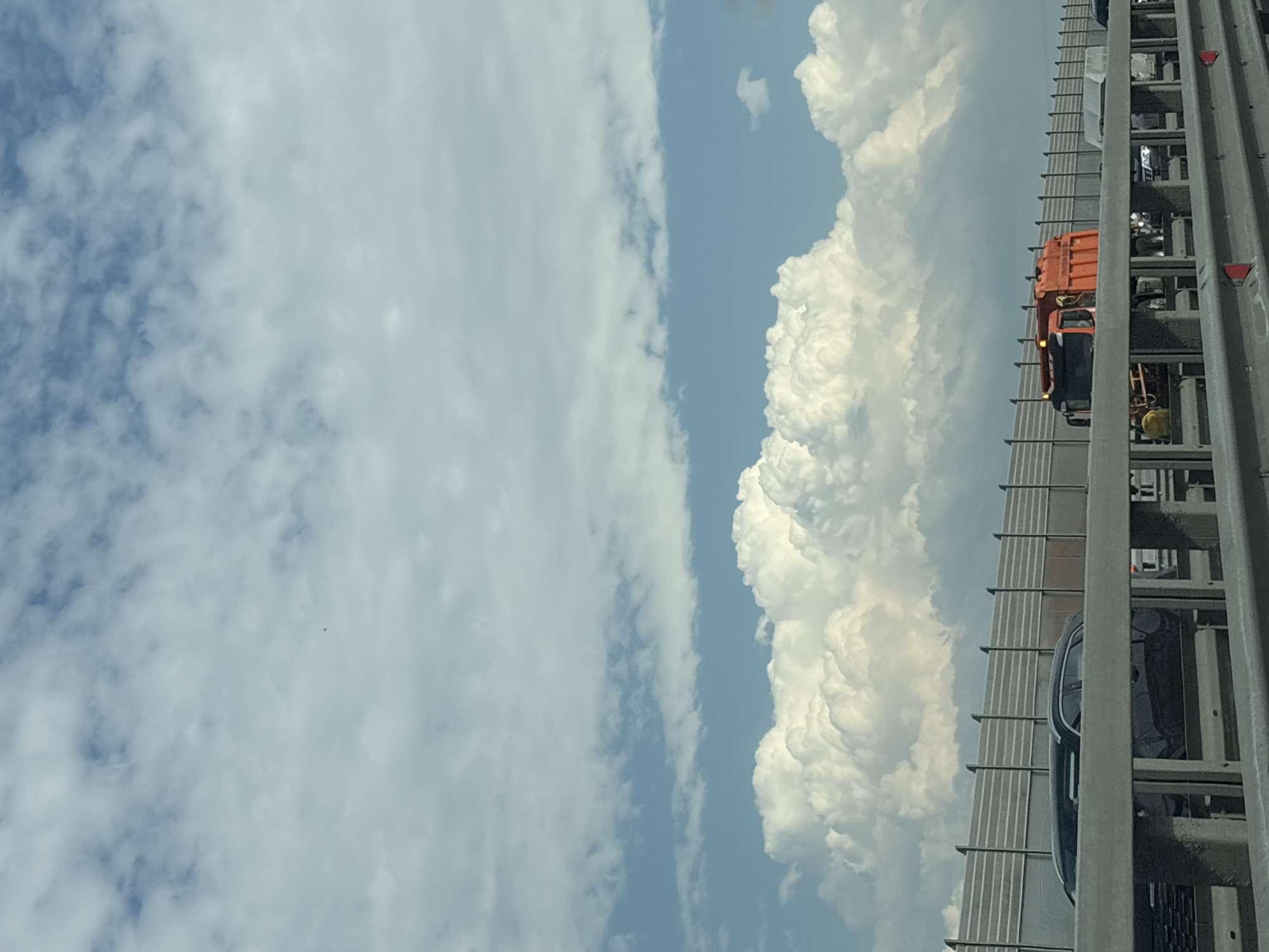 The image presents a view from a vehicle traveling on a highway. To the right, cars and a garbage truck are visible, separated by a guardrail. The sky, predominantly blue, is filled with cumulus clouds. The location is unknown and there is no information to suggest it is in Yukki, Russia. There are no people visible in the image.