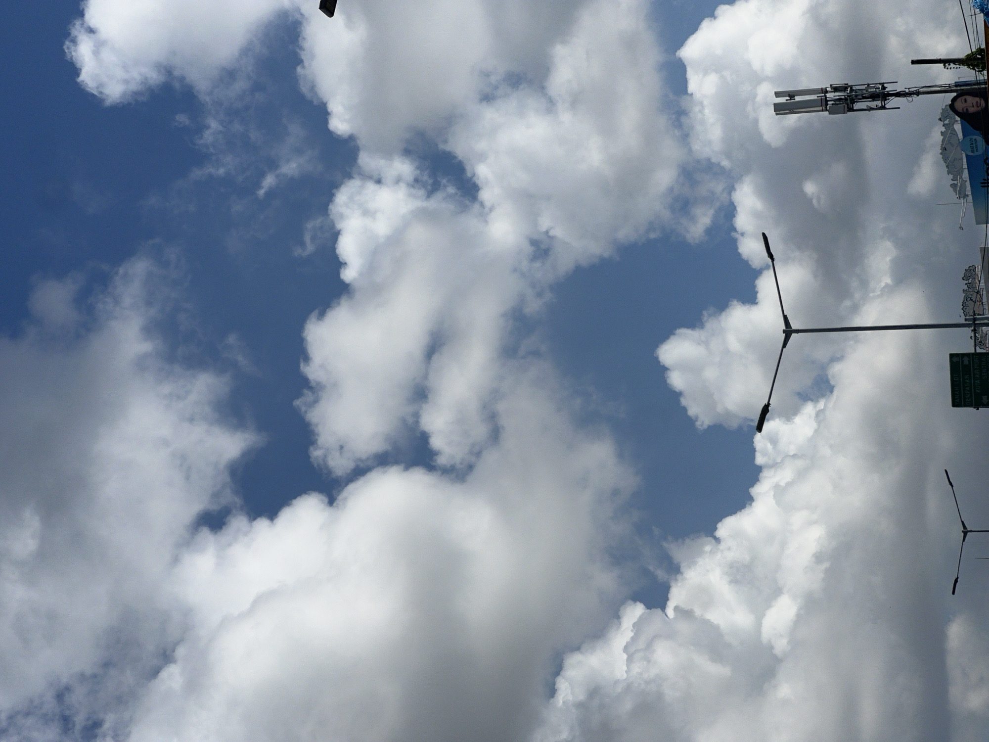 The image captures a cloudy sky over Kuta, Indonesia. In the foreground, several streetlights are visible, with one prominent streetlight in the center extending to the left and right. To the right, a cell tower stands tall with its antennas. Below the cell tower, a woman with dark hair is partially visible, looking upwards. A green directional sign is also present in the lower portion of the image. The sky is filled with large, fluffy white clouds against a backdrop of blue sky. The scene appears to be an outdoor urban setting, likely a street or roadside, under a partly cloudy sky.