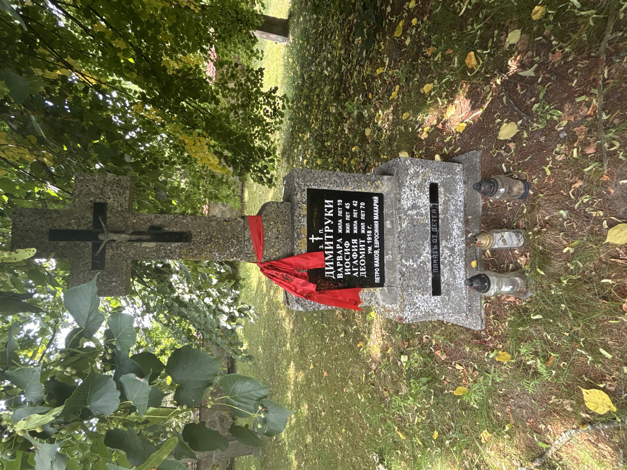 This is a close-up, eye-level shot of a tombstone in the village of Puchły, Poland. The tombstone is made of grey granite with a black plaque in the center. The plaque has text in Cyrillic script, indicating a burial of individuals with the surname "Димитруки" (Dimityuki). The text lists names such as "Варвара" (Varvara), "Иосиф" (Iosif), "Агафия" (Agafiy) and "Деомит" (Deomit) along with their lifespans in years. Below this, it mentions "Петро Иаков, Евфросиния Макарий" (Petro Iakov, Efrosinia Makariy) and a year "1918 г." (1918). A red ribbon is tied around the upper part of the tombstone, and a crucifix is affixed to the top of the stone. In front of the tombstone, three candle holders, two glass and one metal, are placed on the ground amidst fallen leaves and grass. The background shows trees with green and yellowing leaves, and a partially visible stone wall and another tombstone to the right, suggesting it is a cemetery. The lighting is natural, coming from above and to the right, creating some shadows.
