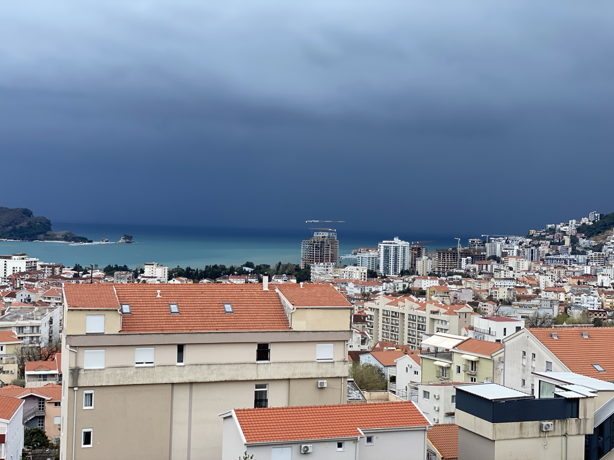 An elevated, wide-angle photograph captures the coastal city of Budva, Montenegro, under a heavily overcast sky. The foreground features several multi-story buildings constructed with light-toned stucco facades in shades of beige and off-white, topped with prominent terracotta-orange tiled roofs. Visible architectural details include rectangular windows, some fitted with white roll-down shutters, and external air conditioning units mounted on walls.
The midground presents a dense urban sprawl, consisting of a blend of older, low-rise structures with similar red tiled roofs and numerous modern, taller apartment complexes. Significant construction activity is evident along the coastal strip, with multiple partially built high-rise structures and several tower cranes visible.
The background displays the Adriatic Sea, exhibiting a turquoise coloration. The sea is bordered by a dark, slate-blue, heavily clouded sky that extends across the upper portion of the frame. A small, vegetated island or peninsula is positioned on the far left within the sea, accompanied by two smaller rocky islets further offshore. No individuals or discernible actions are present within the image.