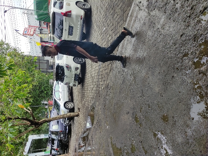 This image captures an outdoor daytime scene in Kecamatan Asembagus, Indonesia, likely an urban or suburban area. The main subject is a young man, possibly of East Asian descent, positioned slightly to the left of the center. He is dressed in a dark short-sleeved collared shirt, dark trousers, and dark shoes. He is captured in a dynamic pose, with his left leg extended straight backward and his right leg bent, giving the impression of him balancing, mid-action, or perhaps playfully attempting a kick or a suspended movement. He is looking towards the camera with a gentle smile.
The setting consists of a paved ground surface on the left, possibly made of bricks or blocks, where several cars are parked. The right side of the ground is a rougher, concrete or asphalt-like texture, heavily covered in mud and standing water, suggesting recent rainfall or poor drainage. In the background, multiple white cars, resembling compact SUVs or minivans, are parked. Further behind them, green trees and foliage are visible, along with parts of buildings and what appears to be a commercial sign with red and white elements, though no specific text is discernible. The overall lighting suggests it is bright daylight. The scene appears to be an everyday moment, perhaps on a street or parking area adjacent to shops or residences, marked by the wet and muddy ground conditions.