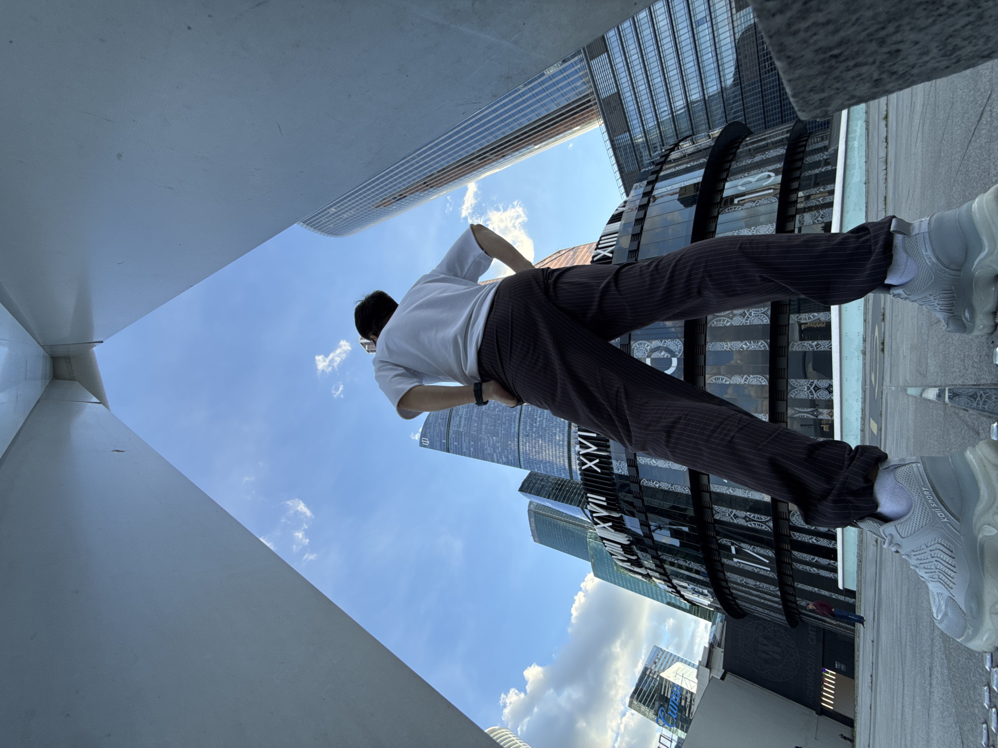 A low-angle, full-body shot shows a person standing with their back to the camera, looking up at the skyscrapers of Moscow City. The person is wearing a white t-shirt, dark pinstriped trousers, and grey sneakers. The background features several modern glass buildings, including the Neva Towers and the Eurasia Tower, under a partly cloudy blue sky. The foreground is a concrete platform with a visible metal grate. The perspective frames the person within a triangular architectural element.