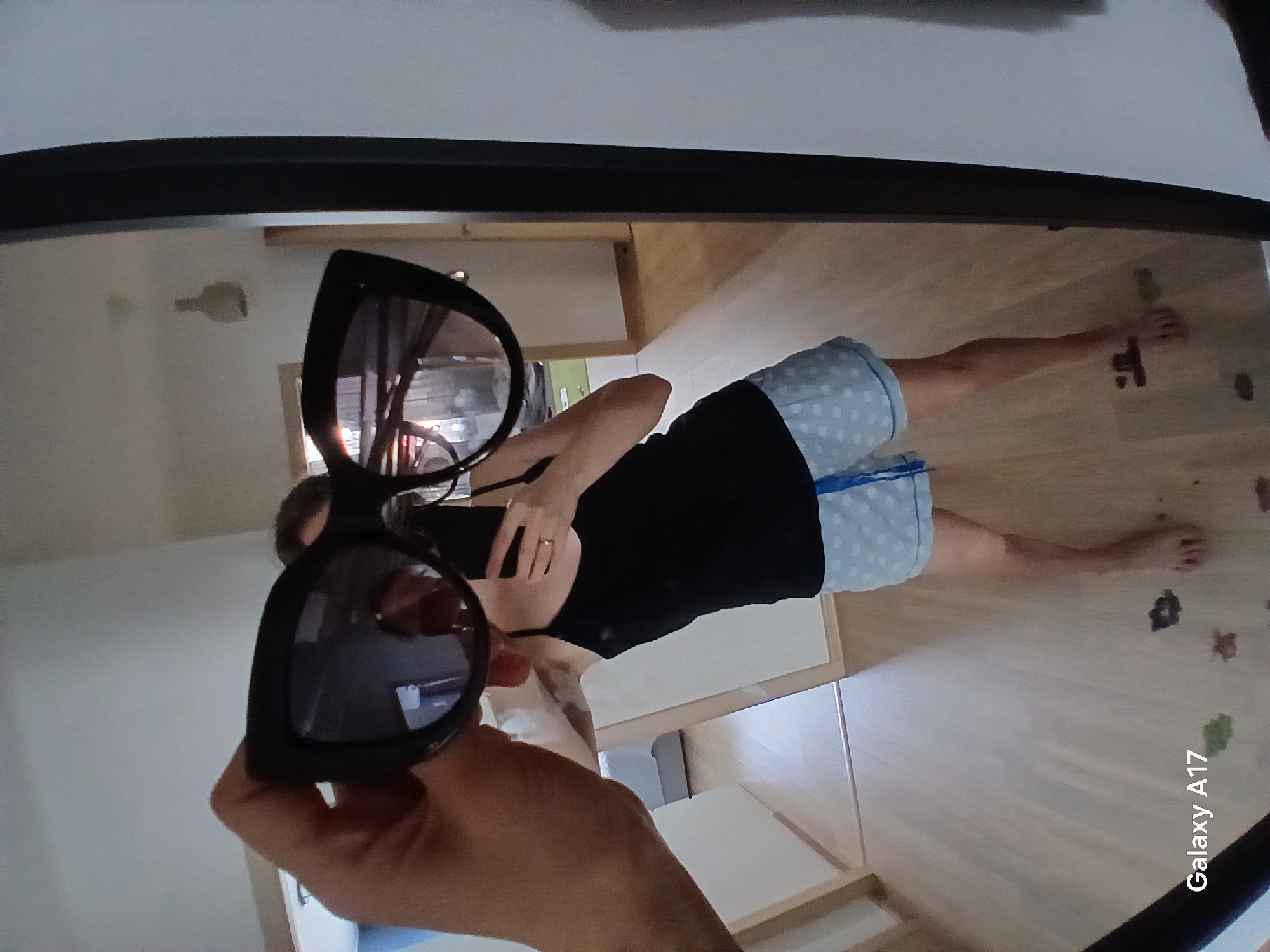 A person is reflected in a framed mirror, holding black sunglasses up to the mirror. The person wears a black tank top and blue and white polka dot shorts. The reflection shows a room with light-colored walls and flooring. In the background, a smartphone is partially visible in the person's hand. The words "Galaxy A17" are visible in the bottom right corner. The location or the city of Yekaterinburg, Russia, cannot be determined from the image.