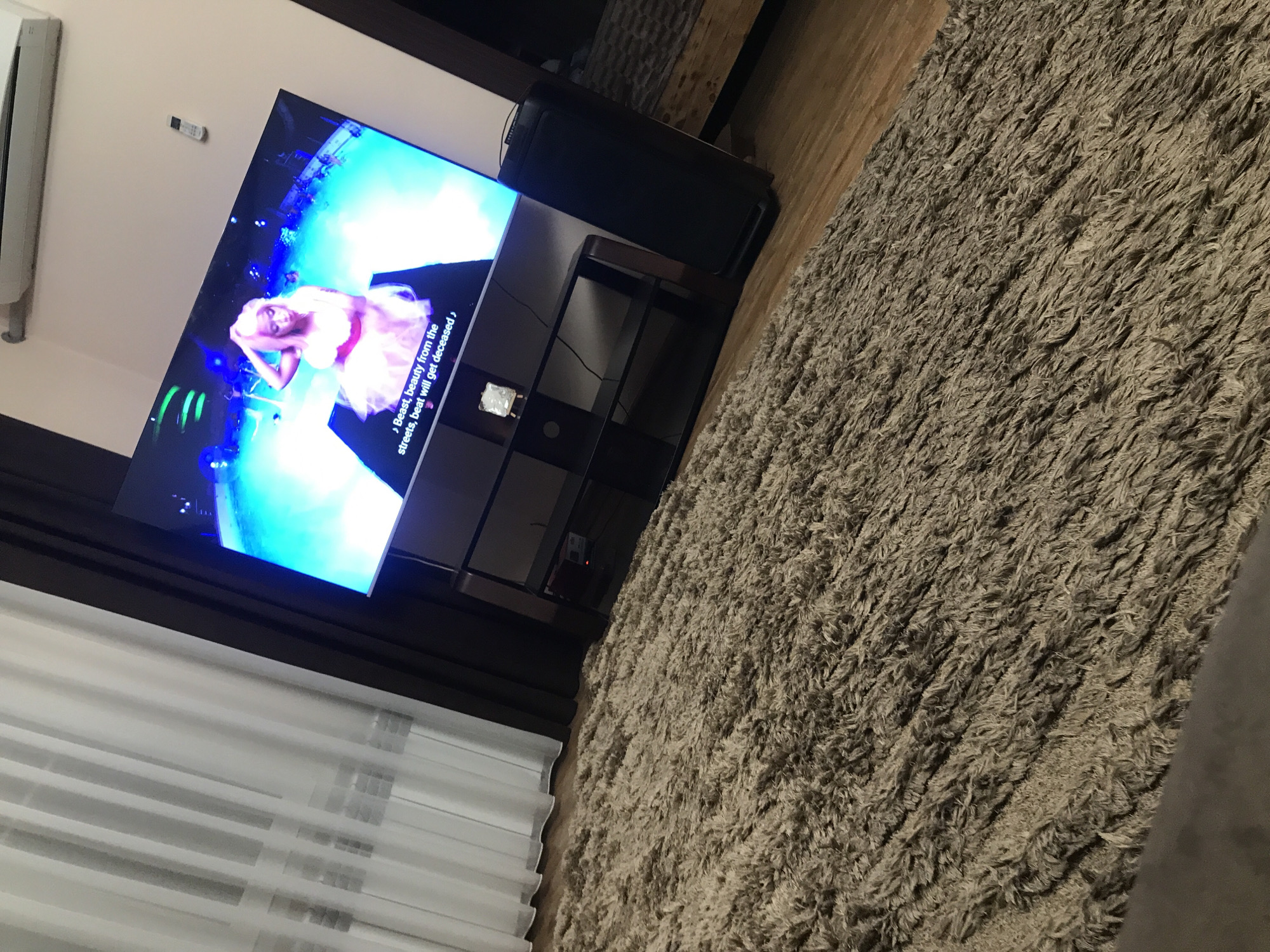 The image shows a living room interior. A large flat-screen TV displays a scene featuring a person in a pink outfit, possibly performing on stage. The TV is mounted on a dark-colored stand with shelves. To the right of the TV stand is a speaker. A large, shaggy rug covers a significant portion of the floor. Light-colored curtains hang near a window. The setting suggests a residence, but the specific location within Yerevan, Armenia, cannot be determined from this single image. There are no apparent people in the photograph beyond what is displayed on the TV. The action is limited to what is visible on the TV screen. The environment is a furnished living room.