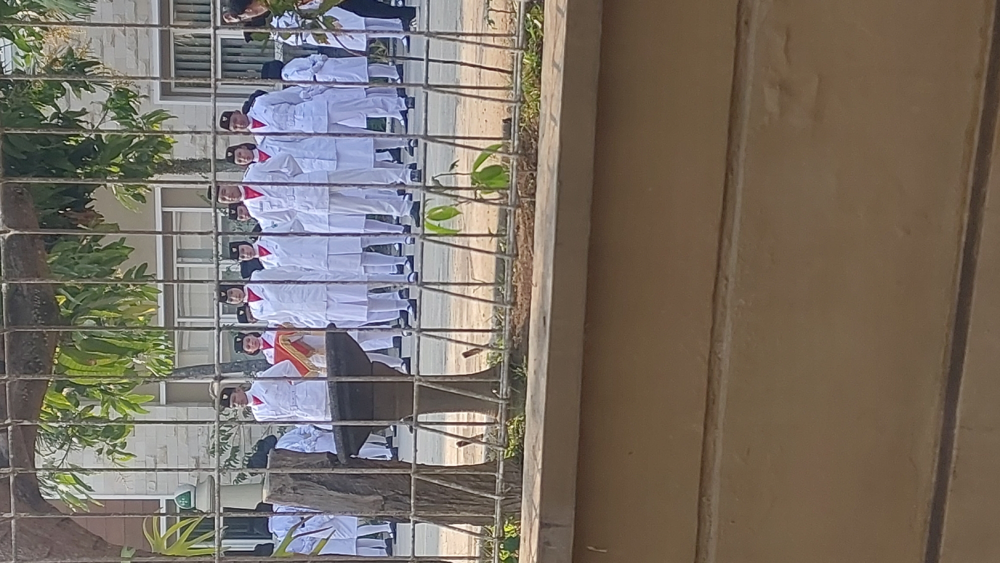 In Malang, Indonesia, a group of uniformed students stands in formation behind a metal fence. The students, all wearing white uniforms with red accents and black caps, appear to be participating in an outdoor ceremony or parade. The building in the background has light-colored stone walls and windows with blinds. The foreground of the image is dominated by the fence and a low, tan-colored wall. The environment suggests a public or institutional setting.