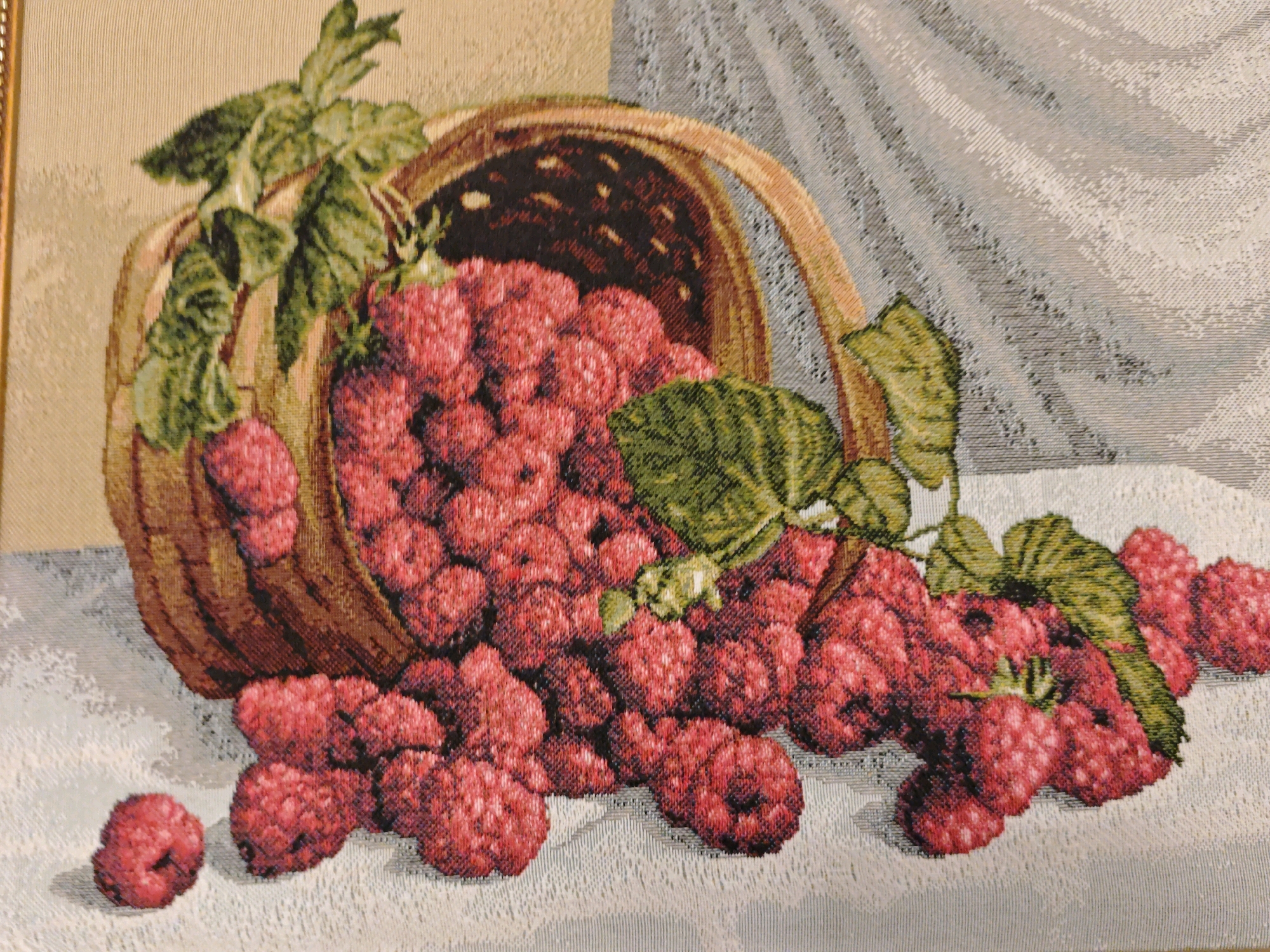 This is a tapestry depicting a still life of raspberries. A rustic woven basket, tilted on its side, is overflowing with ripe red raspberries and green leaves. Several raspberries have spilled out of the basket and are scattered on a textured surface that appears to be a tablecloth. The background of the tapestry is abstract, with muted tones of beige and gray, suggesting fabric drapes or a textured wall. There are no people or animals in the image, and no discernible elements indicating a specific location in Yevpatoriya, Ukraine. The scene is static and focuses solely on the arrangement of the fruit and basket.
