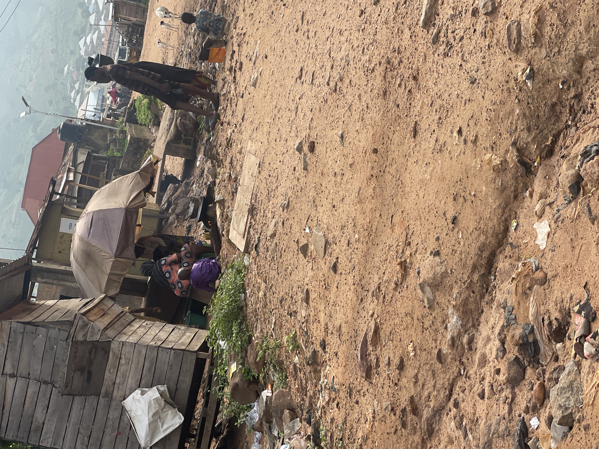 The image depicts a bustling scene in Lokoja, Nigeria, characterized by a dirt road littered with debris, including plastic bags and rocks. To the left, a dilapidated wooden structure stands, with a white bag hanging from its side. In front of the structure, an umbrella shields a group of people, including a child in a purple garment sitting on the ground. To the right, a man in a checkered shirt and dark shorts stands with his back to the camera, carrying a load on his shoulder. Nearby, two dogs are visible, and another child is seated on the ground. In the background, the urban landscape of Lokoja unfolds, with buildings, vehicles, and a street light suggesting the presence of infrastructure. The overall impression is one of a vibrant, yet impoverished, community.