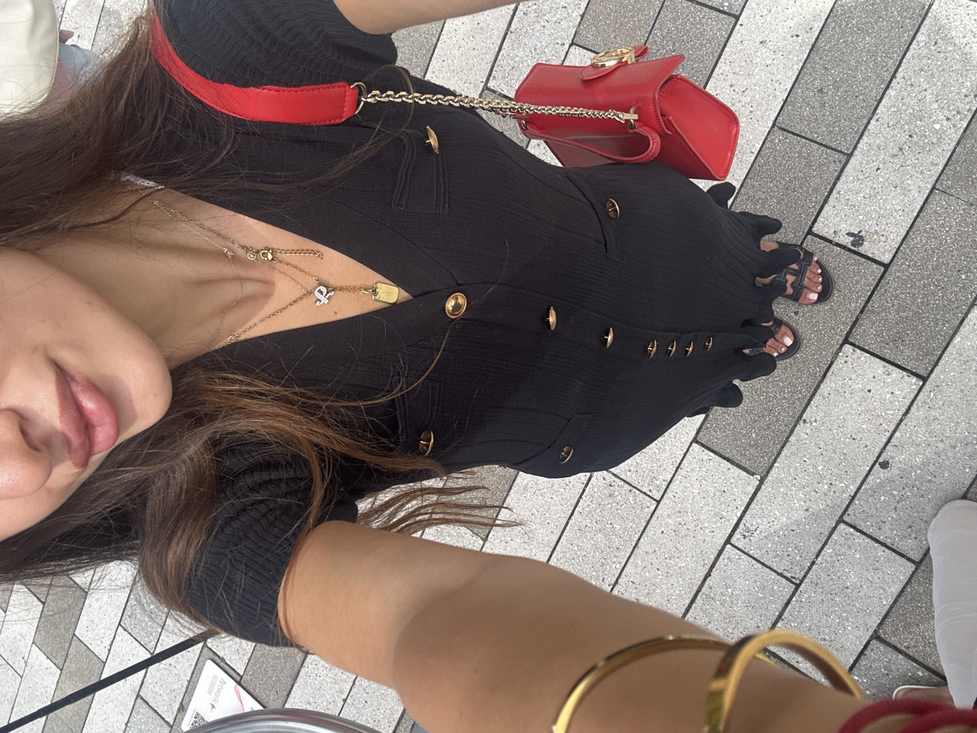 A medium shot captures a woman from the chest down, standing outdoors on a sidewalk paved with light gray and white rectangular tiles. She is wearing a black, ribbed, V-neck dress with short sleeves and gold buttons down the front. The dress has two gold-buttoned pockets on the chest. She is also wearing black sandals with white toenail polish. A gold bracelet is visible on her left wrist, and a bright red handbag with a gold chain strap is slung over her right shoulder. She is also wearing a necklace with multiple gold pendants, one of which is a small, gold-toned letter "Y" with diamonds. Her hair is long and brown. The image provides a clear view of the woman's outfit and accessories against the backdrop of the tiled sidewalk. The location is identified as the city of Aventura, United States of America, though no specific landmarks are visible in the frame to confirm this.