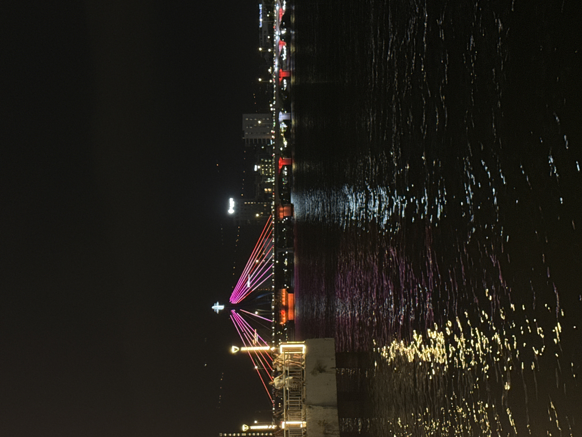 The image shows the Dragon Bridge in Da Nang, Vietnam at night. The bridge is illuminated with colorful lights, predominantly pink and red. The bridge spans across a body of water, which reflects the lights from the bridge and the surrounding city. On the left side of the image, there is a walkway or pier with a railing and several people standing and observing the bridge. The water is dark, with shimmering reflections of lights. In the background, the city skyline is visible with other illuminated buildings and lights. The sky is dark and clear, indicating nighttime.