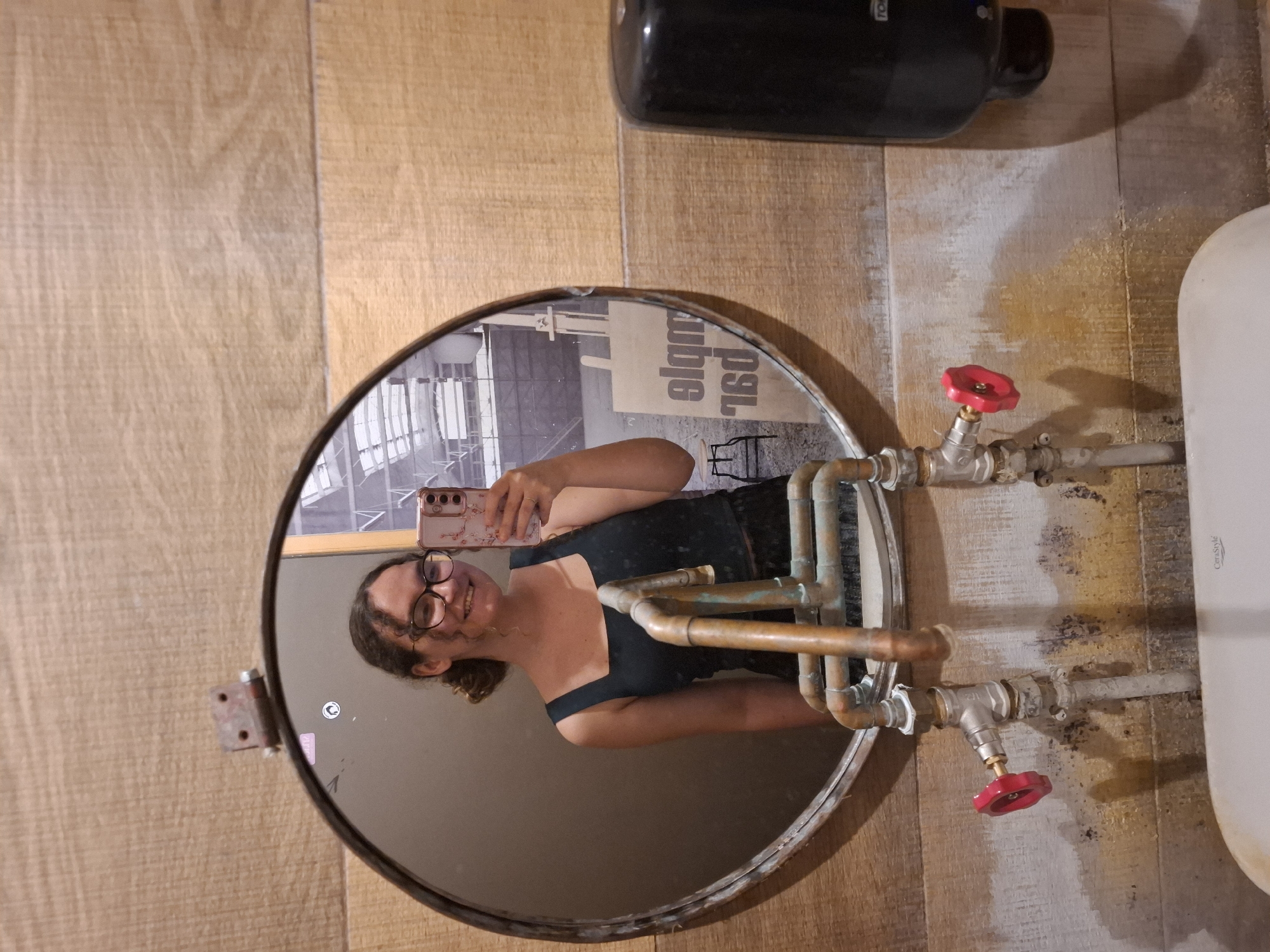 A medium shot captures a young woman in a restroom, holding a smartphone up to take a mirror selfie. The woman has curly brown hair pulled back, is wearing glasses, and a dark tank top. She is smiling at her reflection, which is visible in a large, round, metal-framed mirror. Behind her, visible in the mirror, is a large, open space with what appears to be industrial elements, including large windows or skylights and what looks like a construction site. In the foreground, a sink with exposed copper pipes and red-handled faucet valves is visible. To the right of the mirror is a black soap dispenser mounted on a wood-paneled wall. The overall environment appears rustic, with a mix of wood paneling and what seems to be a tiled wall near the sink.