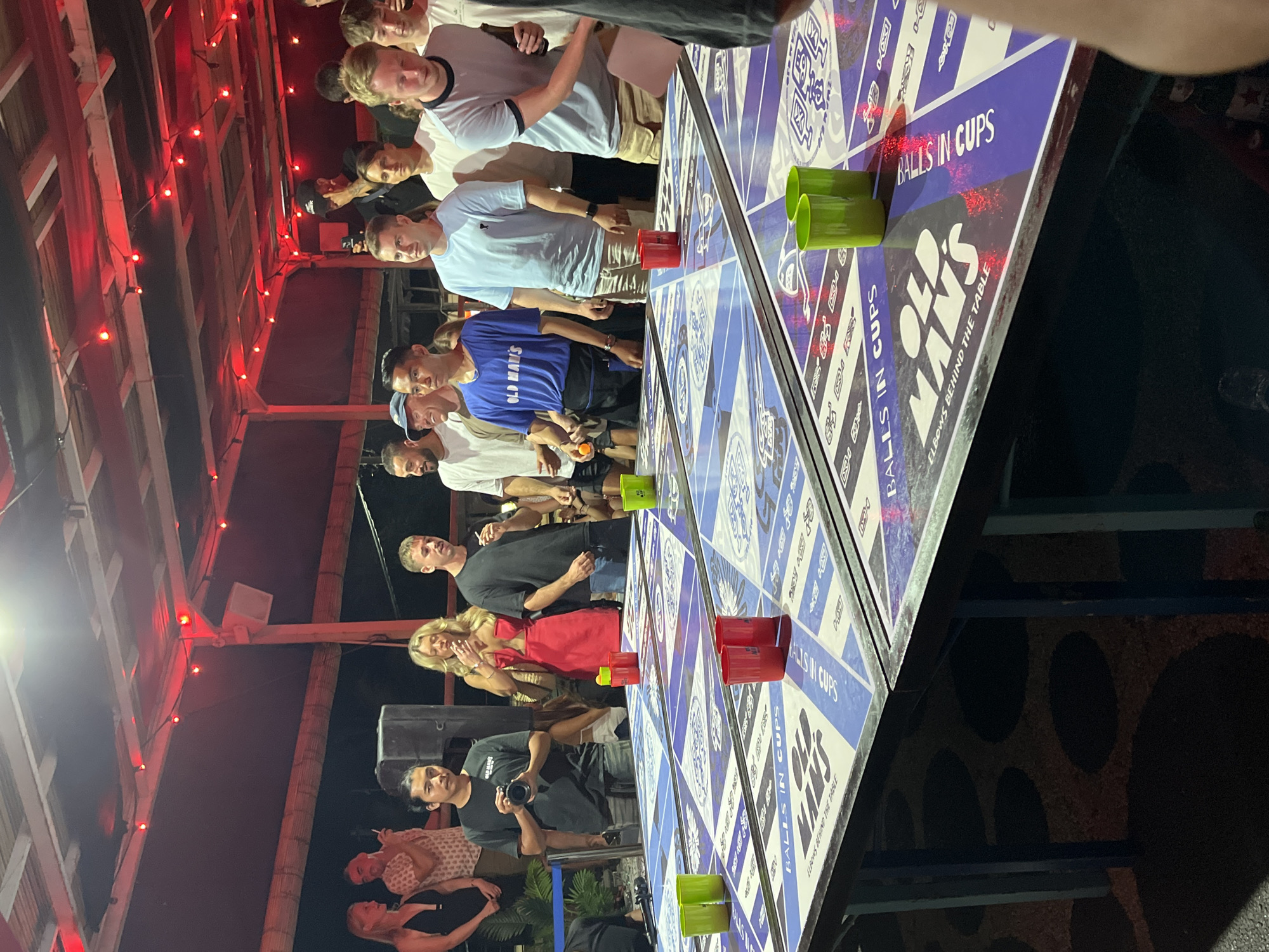 In North Kuta, Indonesia, a group of young adults are gathered around tables set up for a game of beer pong. Red and green cups are arranged on the tables, which are decorated with "Old Man's" branding and the phrase "Balls in Cups." Some individuals are holding drinks or cigarettes. The scene is illuminated by red string lights hanging from a thatched roof structure, suggesting an outdoor or semi-outdoor venue. The participants are dressed casually, and the atmosphere appears lively, indicative of a social gathering or party.