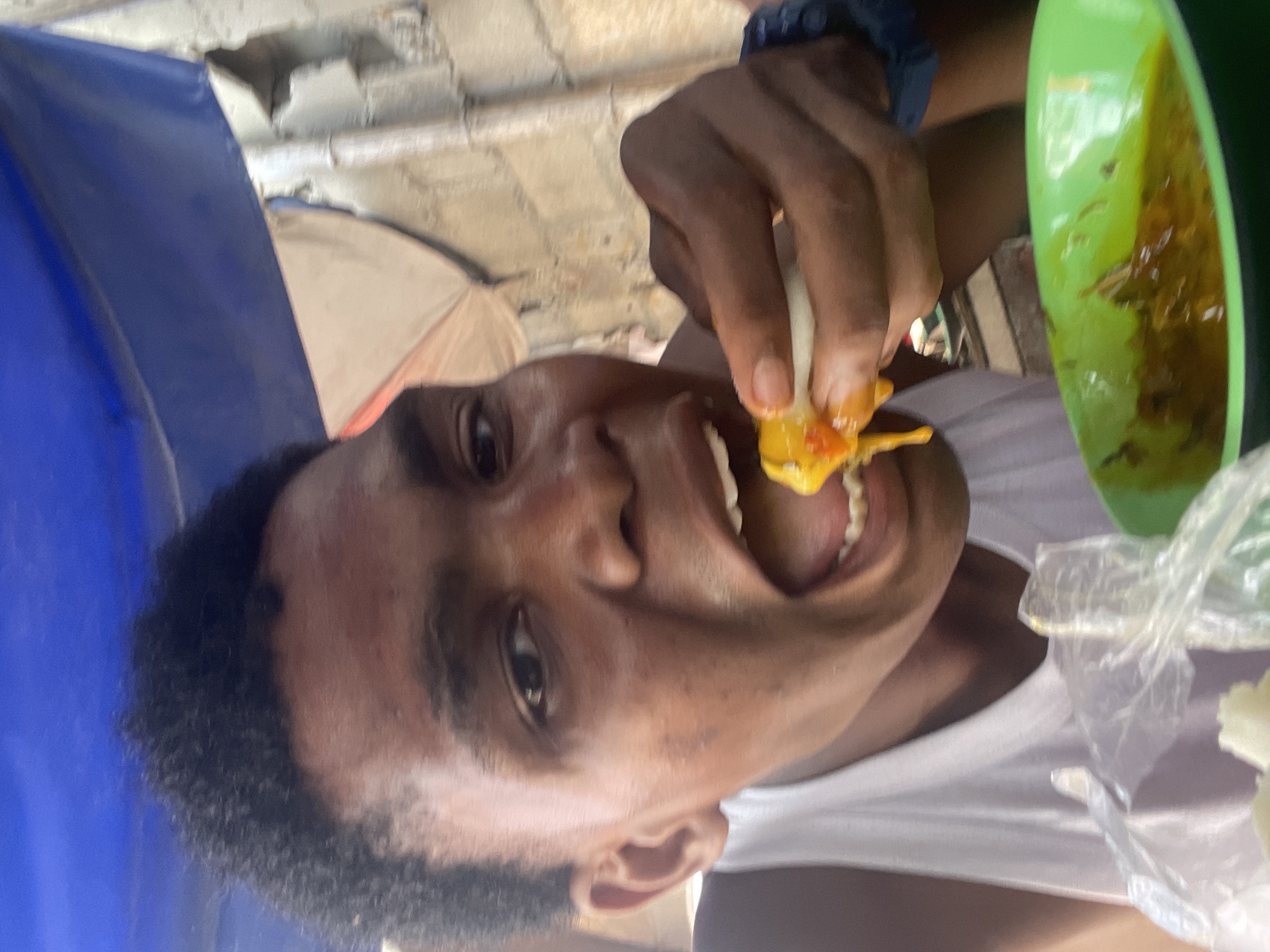 A man with dark skin, short, dark hair, and wearing a white t-shirt, is holding a food item with a white exterior and a bright orange/yellow interior near his open mouth. He appears to be about to take a bite. He is also wearing a blue bracelet. Next to him is a green bowl containing a yellow/brown sauce. A clear plastic bag is visible nearby. The scene appears to be outdoors, potentially under some type of makeshift shelter or tent, as blue and white fabric can be seen in the background along with a cinder block structure. The location is not specified in the image. Therefore it is impossible to determine that this picture was taken in Lekki, Nigeria.