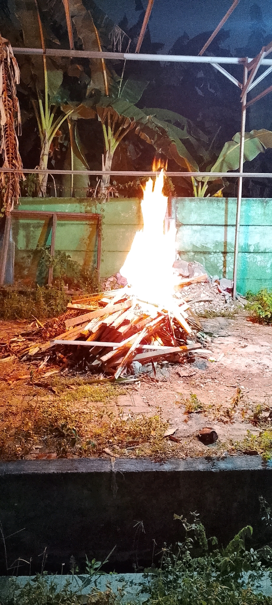 A bonfire burns brightly in front of a teal-colored concrete wall. The fire is built from a pile of wooden planks and other debris, and the flames are a vibrant orange and white. Behind the wall, large green leaves of what appear to be banana trees are visible. Above the trees, a metal structure, possibly a canopy frame, is partially visible against a dark sky. In the foreground, dry grass and some weeds grow around a concrete border. The scene appears to be an outdoor area in Satriamekar, Indonesia, at night.