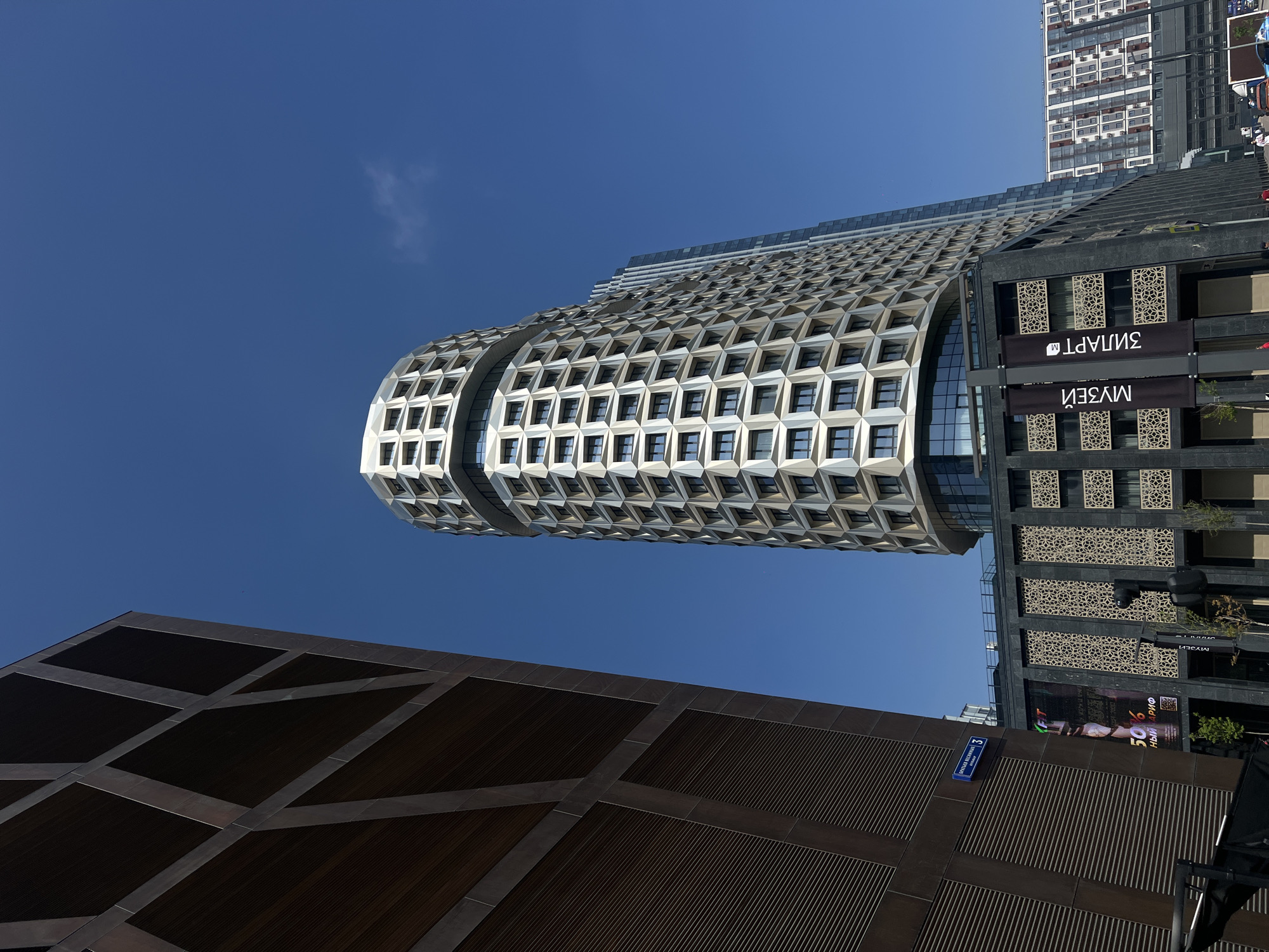 The image shows a low-angle view of modern buildings against a clear blue sky. A distinctive architectural feature is a curvilinear building clad in a repeating geometric pattern of windows. Adjacent to it is a building with dark paneling and decorative latticework over windows. In the foreground, there's a darker building with a grid-like pattern. A street sign is visible on the bottom right. Based on the signage in Cyrillic ("MUSEI") and the address plate visible ("ул. Ерохинa, 3"), it is highly likely the scene is in Moscow, Russia. The buildings appear to be contemporary designs, suggesting an urban environment. There are no people visible in the immediate foreground, but there could be some visible in the reflection.