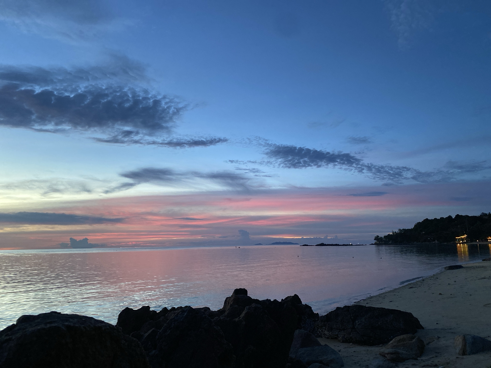 This is a photograph of a tranquil beach scene at dusk on Koh Phangan, Thailand. The sky is a gradient of deep blue at the top, transitioning to hues of pink and orange from the setting sun. Wispy clouds are scattered across the sky, some appearing dark and textured, others light and feathery. The ocean is calm, with gentle ripples reflecting the colors of the sky, creating a soft, diffused light. In the foreground, dark, rough-textured rocks are prominent, some partially submerged in the shallow water. To the right, a sandy beach stretches out, with a faint shoreline where small waves are breaking. Further in the distance, along the coastline, a line of dark green trees is visible, with a few illuminated structures suggesting a small settlement or resort. The overall atmosphere is peaceful and serene, characteristic of a tropical sunset.