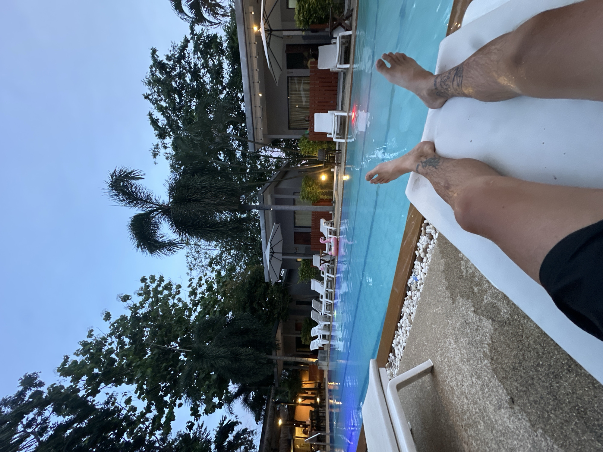 The image shows a person's legs and feet resting on a lounge chair next to a swimming pool. The legs are tanned and have tattoos on the calves. The pool is illuminated with blue lights, and there are several white lounge chairs lined up beside it. In the background, there are palm trees, buildings, and string lights, suggesting a tropical resort setting. A pink inflatable flamingo can be seen floating in the pool. The time of day appears to be dusk or early evening, as the sky is a deep blue and the lights are on. The weather seems to be calm. The scene evokes a sense of relaxation and vacation. The location context is Rawai, Thailand. There is no visible text in the image.