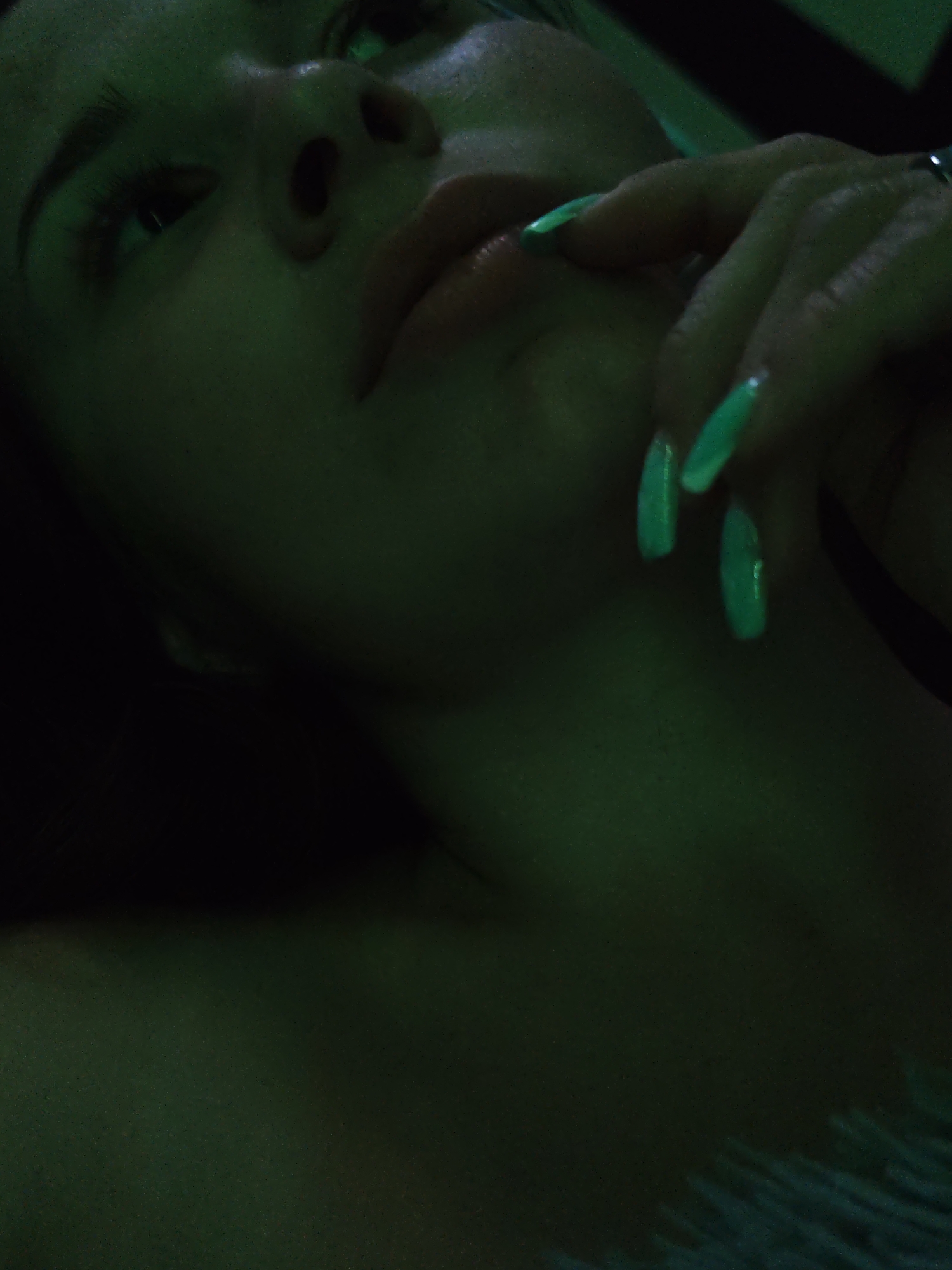 A close-up, low-angle shot of a person with light skin, captured in a green hue. The person's face is prominently displayed, showing their eyes, nose, and lips. Their hand is raised near their mouth, with long, pointed, green-painted nails. The environment is dark, with a few hints of other surfaces. The location is not discernible from the image.