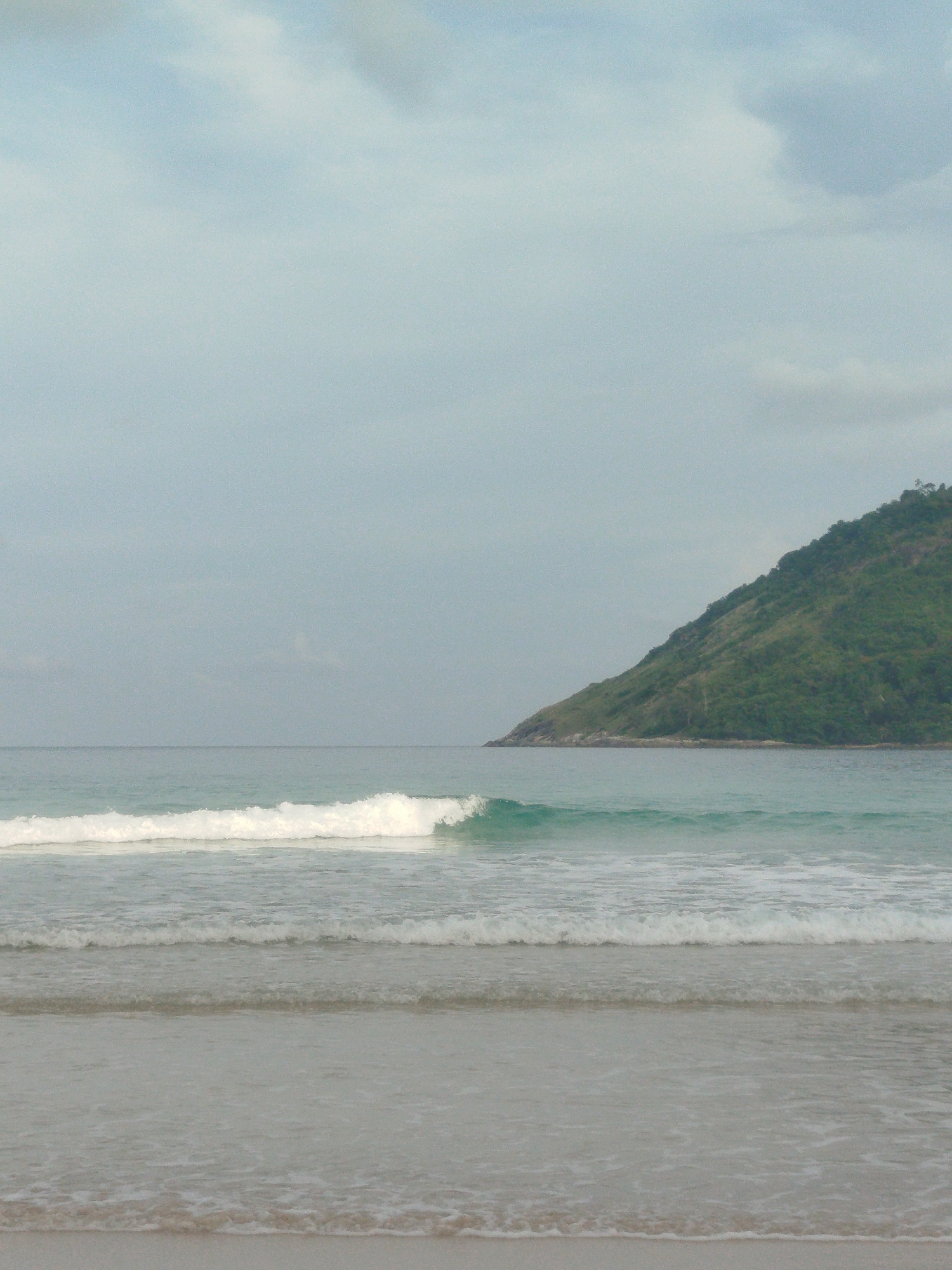 This media file depicts a serene beach scene in Rawai, Thailand. The subjects include the natural elements of the ocean, a sandy shore, and a lush green island in the distance. The primary activity is the gentle lapping of waves on the shore, creating a sense of tranquility.
The time of day appears to be overcast, with soft, diffused light suggesting either early morning or late afternoon. The weather is cloudy, contributing to the subdued and peaceful atmosphere. The presence of a verdant island, characteristic of tropical coastlines, strongly indicates a location in a region like Thailand. The waves are moderate, with white foam indicating they are breaking. The water is a cool, muted turquoise. The sand in the foreground is light-colored and wet from the receding tide.
There are no people or man-made objects visible in the frame, emphasizing the natural beauty of the location. The overall emotion conveyed is one of calm and natural serenity. No text is visible in the image.