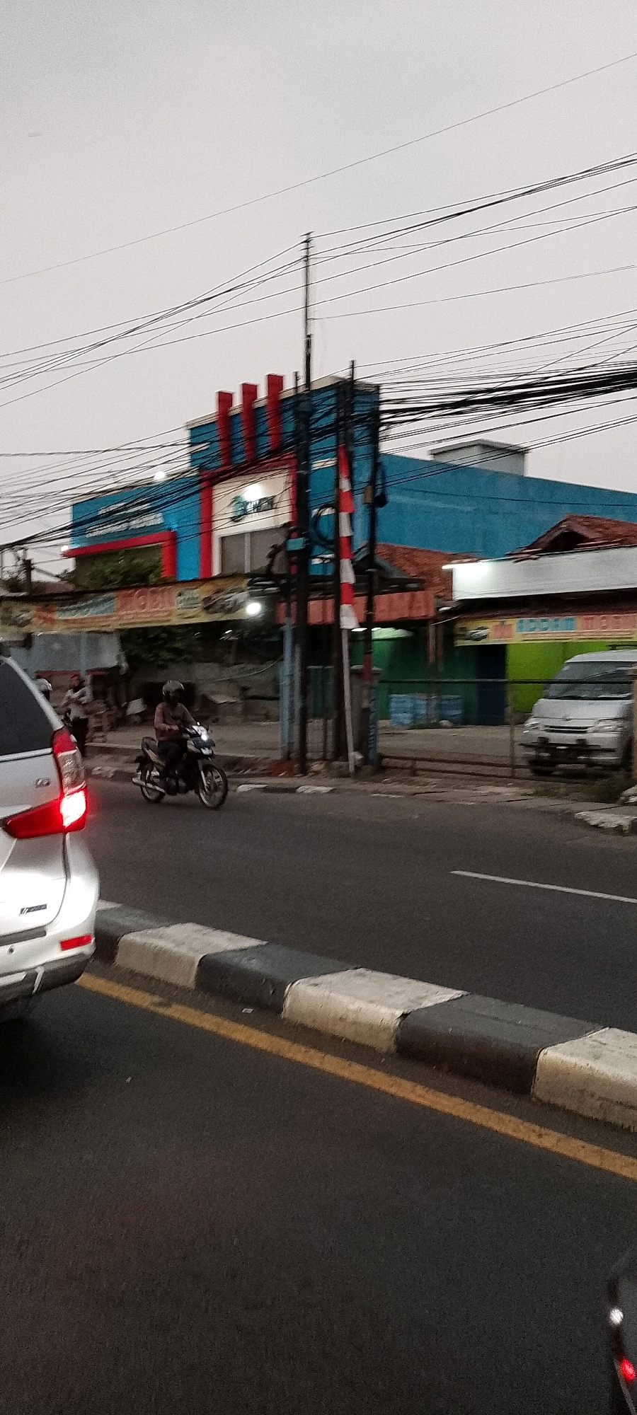 This is a vertical, medium-shot, eye-level, outdoor, daytime photograph taken from the perspective of a vehicle on a street, likely in Depok, Indonesia, given the context. The foreground is dominated by the rear bumper and taillight of a silver car on the left side of the frame, its brake light illuminated. A yellow line and a concrete curb with alternating black and white painted sections mark the edge of the asphalt road.
In the middle ground, a person on a motorcycle, wearing a dark helmet and a patterned shirt, is riding on the road. To the left of the road, a banner with text is visible, partially obscured by a metallic pole. Behind the banner, there are a few people standing near a building.
The background features a two-story building with a blue facade and red decorative elements on the top section. A white circular logo with blue text is present on the building. Several Indonesian flags, red and white, are displayed on poles in front of the building. A white van is parked in front of a green-walled building to the right of the main blue building. A complex network of electrical wires stretches across the sky above the buildings. The sky is overcast and grey.