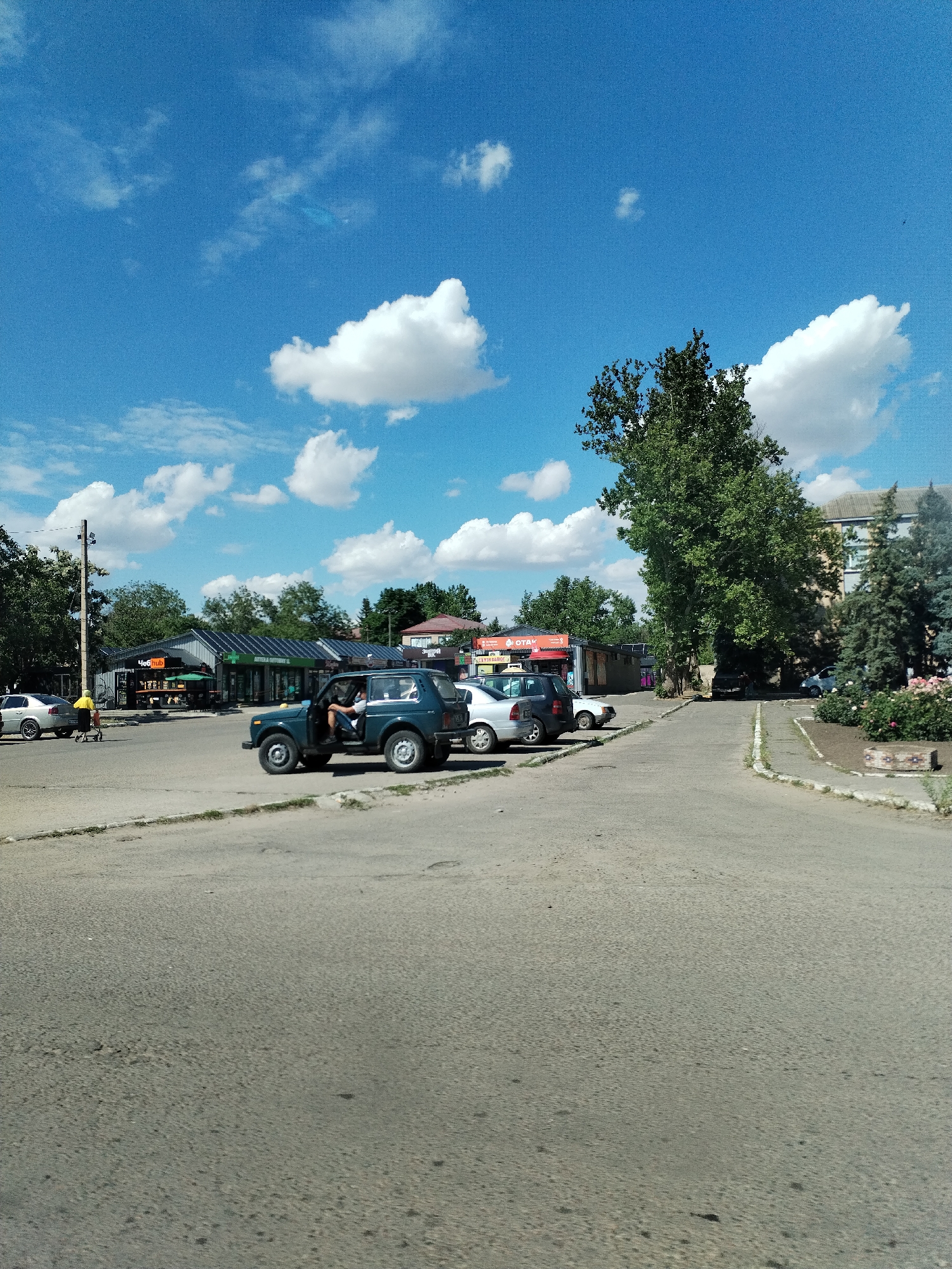 In Nova Odesa, Ukraine, a scene unfolds on a sunny day with a bright blue sky dotted with puffy white clouds. The foreground shows a paved road with some wear and tear. To the left, a silver sedan is parked. Further back, a dark green SUV with its driver's door open is positioned in front of a row of low-rise buildings that appear to be shops or businesses. One of these buildings has a sign that reads "Hub". To the right of the SUV, a white sedan is parked. Beyond these vehicles, the landscape transitions to trees and a multi-story building. A tall, mature tree with dense foliage stands prominently in the middle ground. The area is characterized by a mix of commercial and residential structures, with ample open space and vegetation.