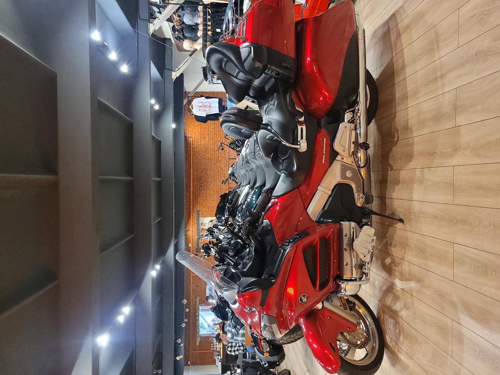 The image shows a motorcycle dealership, likely in Moscow, Russia based on architectural styles. A line of new, predominantly red Honda Gold Wing motorcycles is displayed on a wooden floor, with various models visible. The showroom has a modern design with a dark-painted wall featuring shelving units, illuminated by a string of lights. In the background, there are people browsing clothing items and possibly other motorcycles on display. The setting suggests a retail environment.