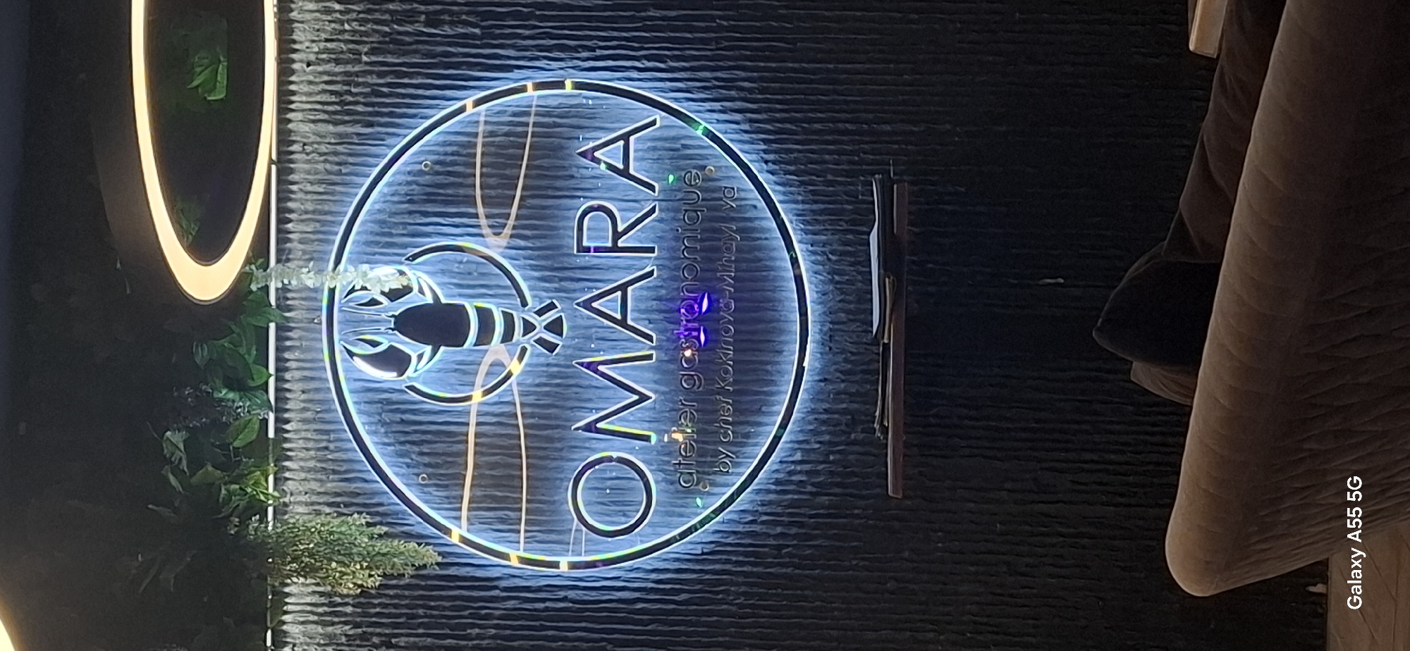 The image is a vertical shot of a restaurant sign and interior. The sign is a circular neon light with a lobster graphic at the top and the words "OMARA" in large letters. Below "OMARA", it reads "atelier gastronomique" and "by chef Kokinova-Mihaylova". The sign is mounted on a textured, dark wall, possibly with vertical slats. Some artificial greenery hangs from above. In the foreground, a portion of a plush sofa is visible, and the bottom left corner shows a "Galaxy A55 5G" watermark. The restaurant appears to be located in Pomorie, Bulgaria.