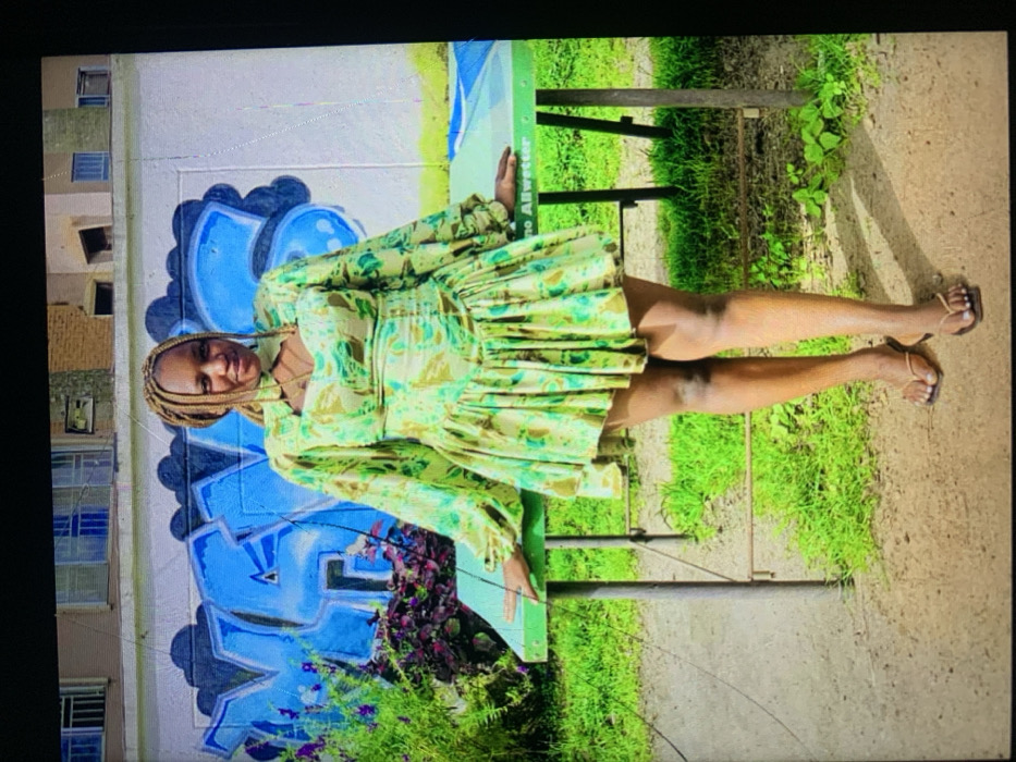 The image depicts a female subject standing outdoors in Jos, Nigeria. She is an adult female with light brown braided hair, facing the camera with a slight smile. She wears a short dress patterned in shades of green, light green, and brown, featuring long, full sleeves and a high neckline with a cutout above the bust. Dark-colored flip-flops are on her feet. Her hands rest on a rectangular light green table with metal legs; the table's surface partially displays the text "no Allwetter." The subject stands on light brown dirt. Behind her and the table is a white wall adorned with large blue graffiti depicting stylized letters. To the left, green foliage and purple flowers are visible. Above the wall, parts of a multi-story building with brown and light-colored sections and windows are present, indicating an outdoor setting under bright daylight.