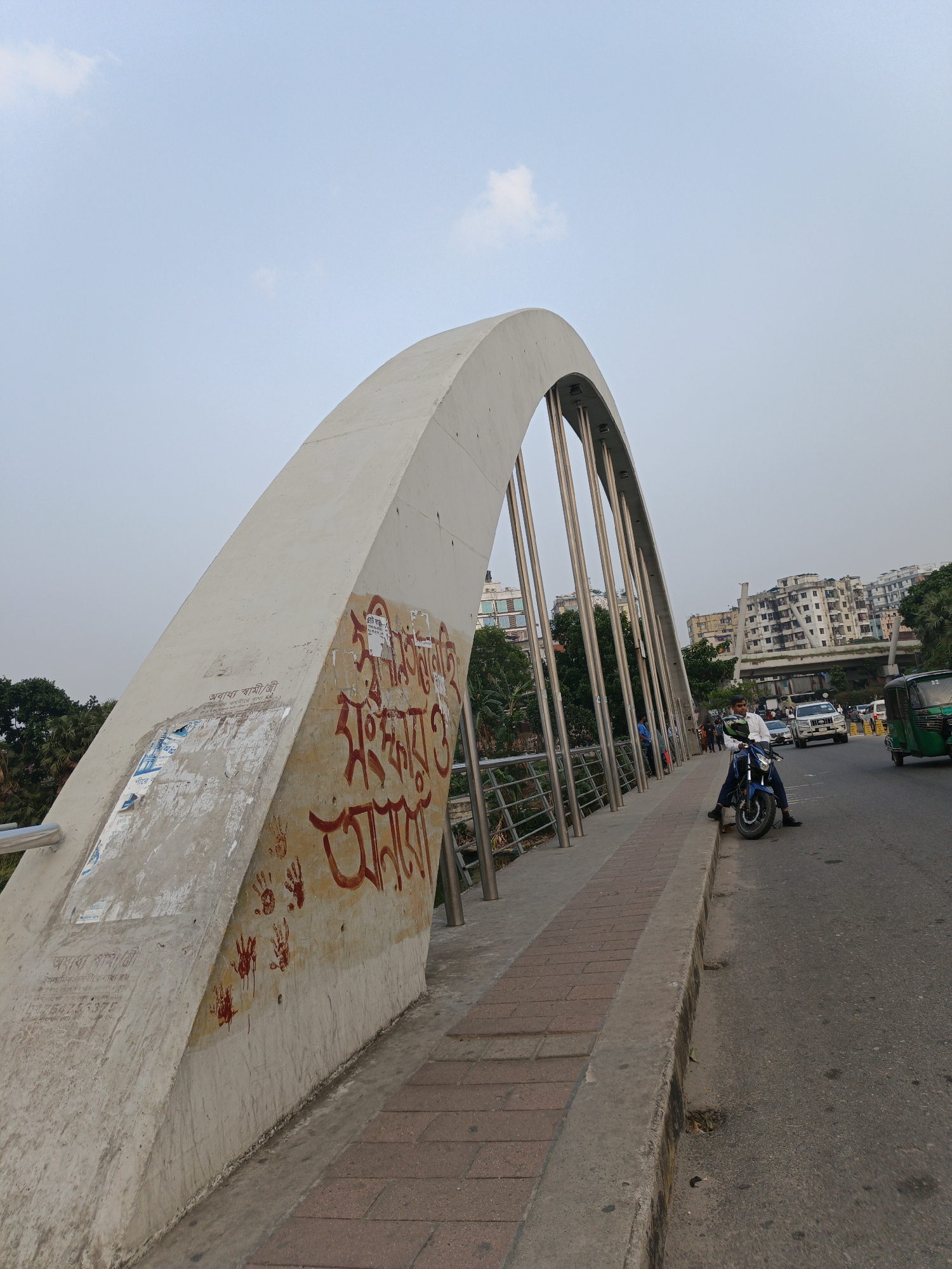 The image captures a scene on a bridge in Dhaka, Bangladesh. The bridge is characterized by a large, white concrete arch. The exterior of the arch is covered with red Bengali script and handprints. A metal railing borders the walkway.
On the right side of the frame, a road is visible, with moving traffic consisting of cars and auto-rickshaws. A man on a blue motorcycle is seen at the edge of the road, alongside the bridge's walkway. In the background, buildings are visible. The sky is light blue with a few scattered clouds.