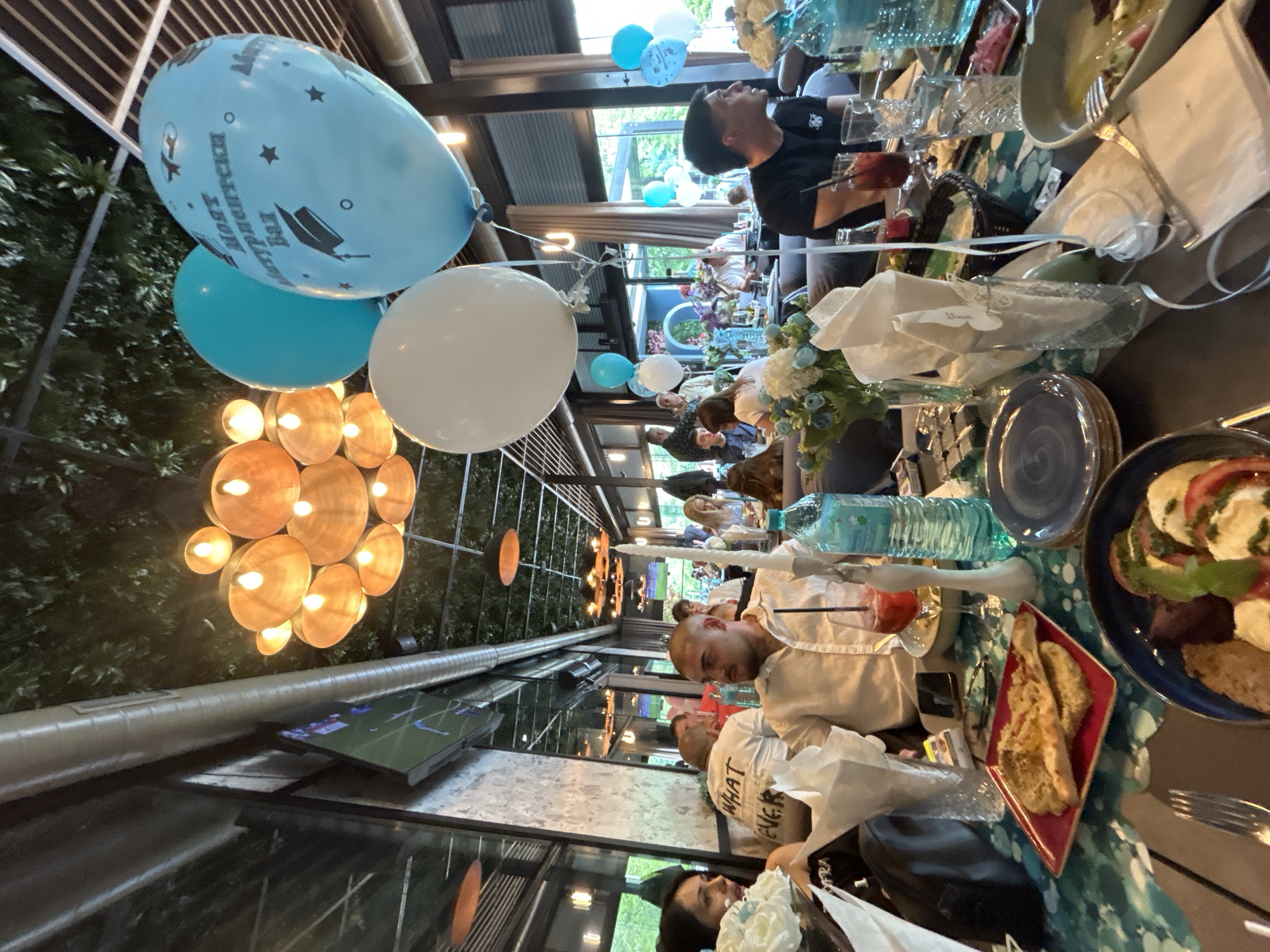 The image shows a gathering of people at a restaurant, possibly a celebration, in Burgas, Bulgaria. Several people are seated at tables, engaged in conversation and dining. There are light blue and white balloons, some with graduation-themed decorations. The tables are set with plates, glasses, water bottles, and food. One person is visible in a black shirt, while others are wearing white shirts. There is a TV screen in the background. The restaurant has a partially open roof and is surrounded by greenery.