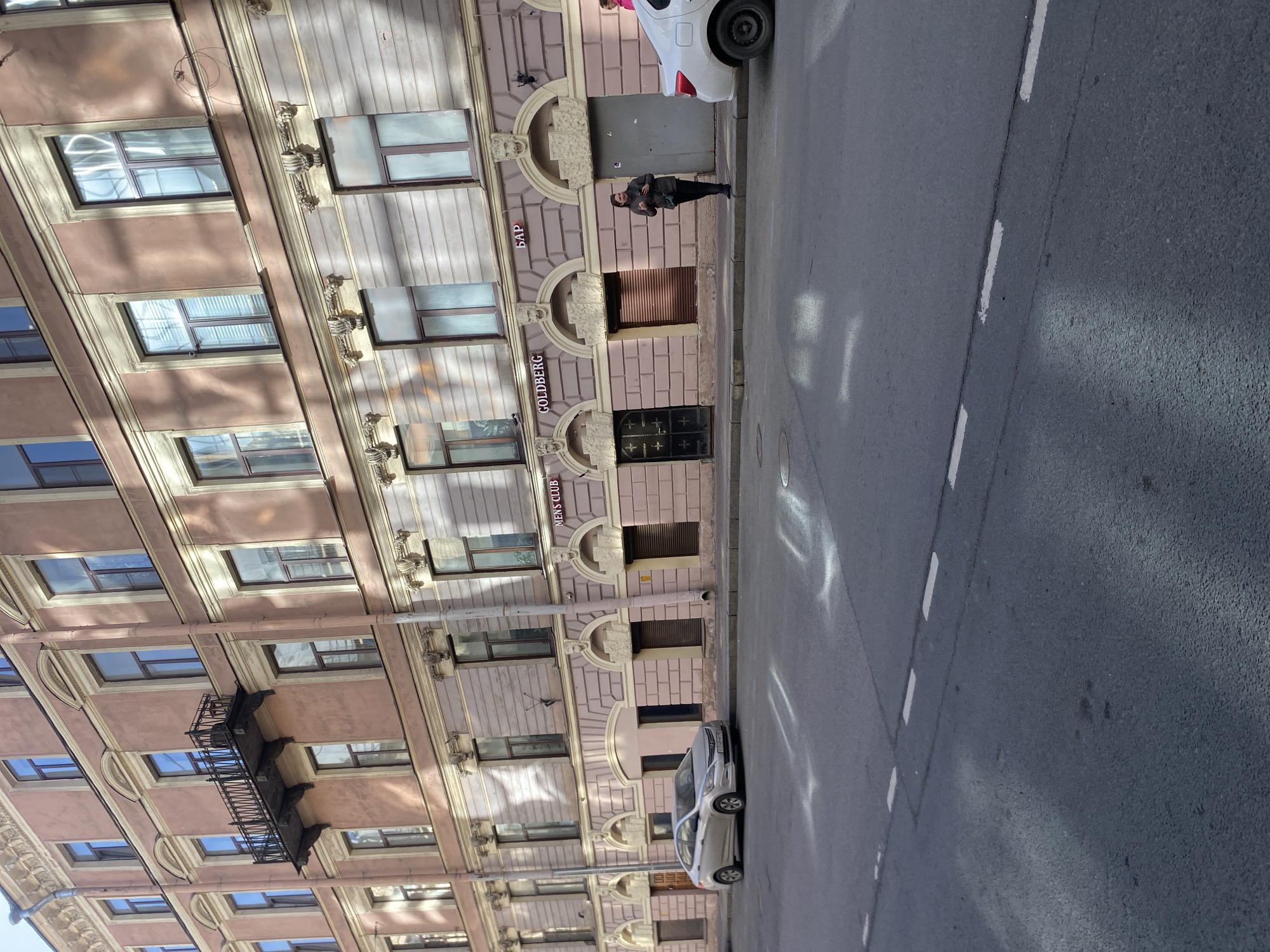 The image depicts Konny Pereulok, 16, Saint Petersburg, Russia, showing a multi-story classical building facade, a street, parked vehicles, and pedestrians under daylight conditions.
The building features a light reddish-brown upper facade with a lighter, pale pinkish-beige ground and first floor, trimmed with cream or yellow architectural details. Numerous rectangular windows with dark frames are visible; some upper windows reflect sunlight and sky, while lower ones appear frosted or shuttered. A single dark wrought-iron balcony is present on the third floor towards the left. Vertical downspouts run along the facade.
The ground floor consists of multiple arched entrances. From left to right, these include a dark recessed doorway, a brown rollup shutter, a dark metal gate with decorative cross patterns (below the "GOLDBERG" sign), another brown rollup shutter, and a plain grey metal door (below the "БАР" sign). Signage on the ground floor includes "MEN'S CLUB" to the left, "GOLDBERG" prominently in the center, and "БАР" (BAR) to the right, all in white or gold letters on dark backgrounds. Decorative lion head reliefs are visible above some ground-floor arches.
The asphalt street in the foreground features white dashed lane markings. Patches of sunlight or reflections are visible on both the street and the building facade.
Two cars are present: a silver Hyundai Solaris sedan (license plate "H748BM 1XX") is parked curb-side on the left, facing right. The rear portion of a white sedan is parked curb-side on the right, facing left.
One woman stands on the sidewalk near the white car, between it and the grey metal door. She wears a dark jacket, dark pants, and dark boots, looking down at a dark object held in her hands. A partial view of another person in a pink jacket is visible further right, behind the white car. Strong diagonal shadows are cast across the upper part of the building facade from left to right.