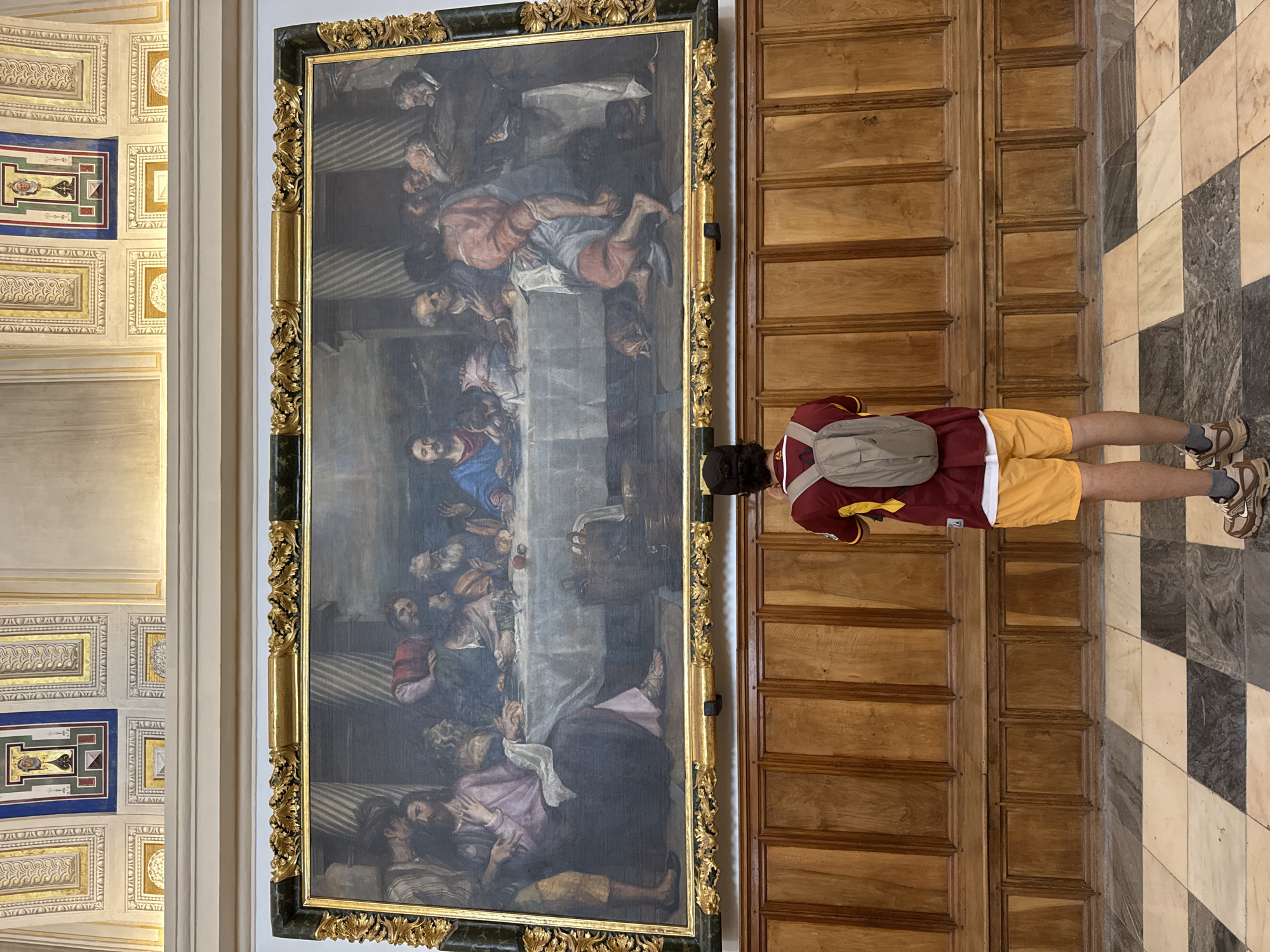 In the Hall of the Kings within the Royal Site of San Lorenzo de El Escorial, Spain, a person stands before a large oil painting depicting "The Last Supper." The painting is framed by an ornate gold frame and hangs on a wall adorned with wooden paneling and benches below. The floor is a checkerboard pattern of light and dark stone tiles. The person is wearing a maroon and yellow shirt, yellow shorts, a grey backpack, and sneakers. They are facing away from the camera, looking intently at the artwork, which features Jesus and his disciples gathered around a table laden with food. The scene is illuminated by natural light, casting soft shadows. The ceiling above the painting is decorated with intricate stucco work.