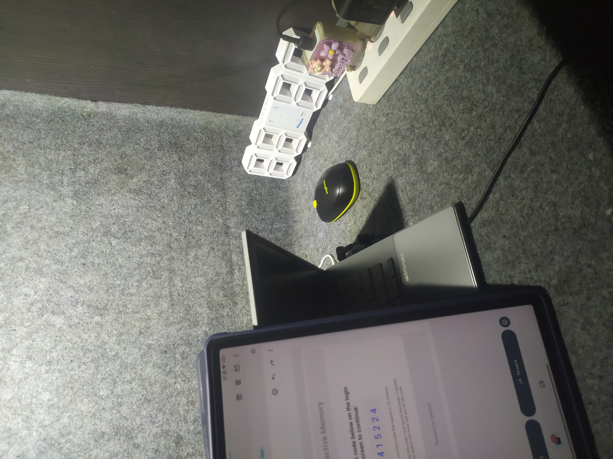 The image shows a dark, textured surface, likely a table or floor covered in grey felt-like material. On this surface, several electronic devices are arranged. In the foreground, a tablet is displaying a screen with text, including a code "415224". To the right of the tablet, a partially opened laptop is visible. Further to the right, a black and yellow Logitech mouse sits near a white digital clock displaying the time. Behind the clock is a power strip with several plugs and a small decorative item featuring a cartoon cat. A black cable snakes across the surface. The overall lighting suggests an indoor setting, possibly a home office or study area. There is no indication of the location being in Kragilan, Indonesia.