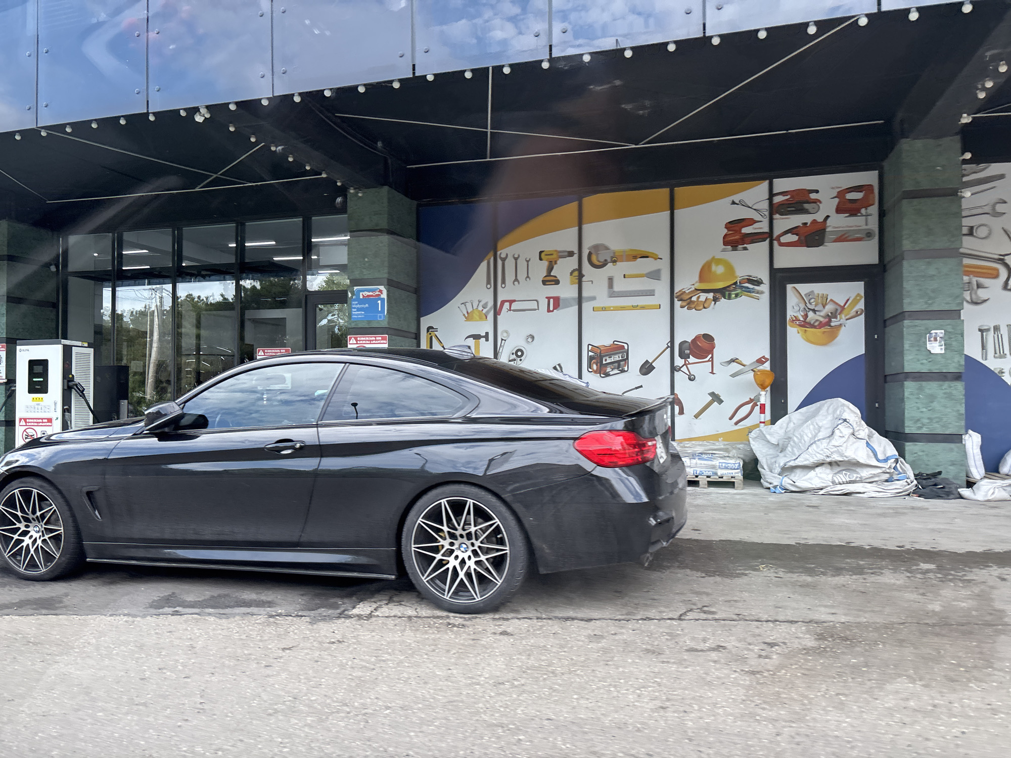 A black BMW 4 series coupe is parked in front of a building with a large window display featuring images of tools. The car is positioned on a concrete surface, with a white electric vehicle charging station to its left. The building's facade is modern, with a black overhang and a glass front. The window display shows various tools such as drills, saws, hammers, and wrenches. To the right of the car, there are large white sacks, some stacked on pallets, and some loose. The scene appears to be a retail or service area, possibly a hardware store or an automotive service center. The environment suggests an outdoor setting, with a cloudy sky visible in the reflection of the building's glass. The overall impression is a commercial area with a focus on tools and equipment.