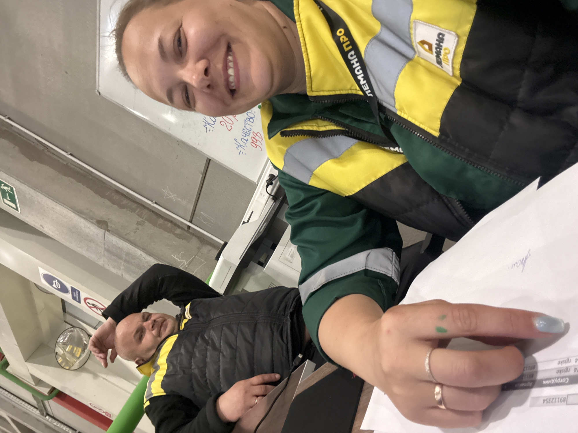The image depicts a selfie of a smiling woman in a yellow and green work jacket, likely indoors. She is holding papers with handwritten notes and printed text. A man in a similar black and yellow jacket is lying in the background. The image includes a whiteboard with writing on it. There is text in the image related to "LEMANA PRO" and some Russian words. The location in Lupanovo, Russia is not discernible from the image.