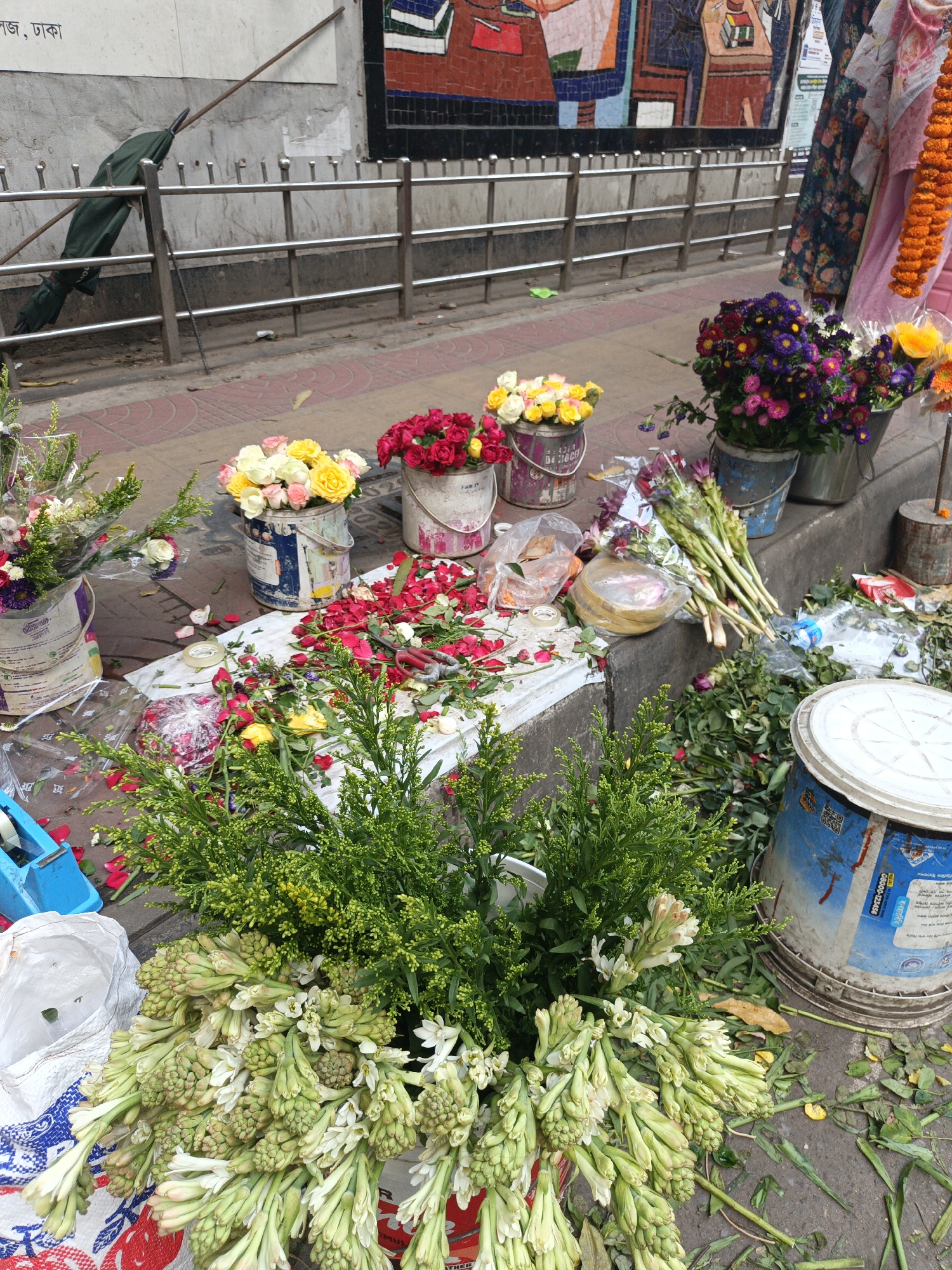 The image depicts a flower stall on a sidewalk in Dhaka, Bangladesh. Buckets filled with flowers in various colors (red, yellow, white, purple) and types (roses, tuberoses) are arranged along the curb. Loose flower petals are scattered on a white sheet laid out on the ground. A silver railing runs along the edge of the sidewalk. An umbrella leans against the railing in the background. A mosaic mural adorns the wall behind the railing. A woman with a marigold garland is standing in the background.