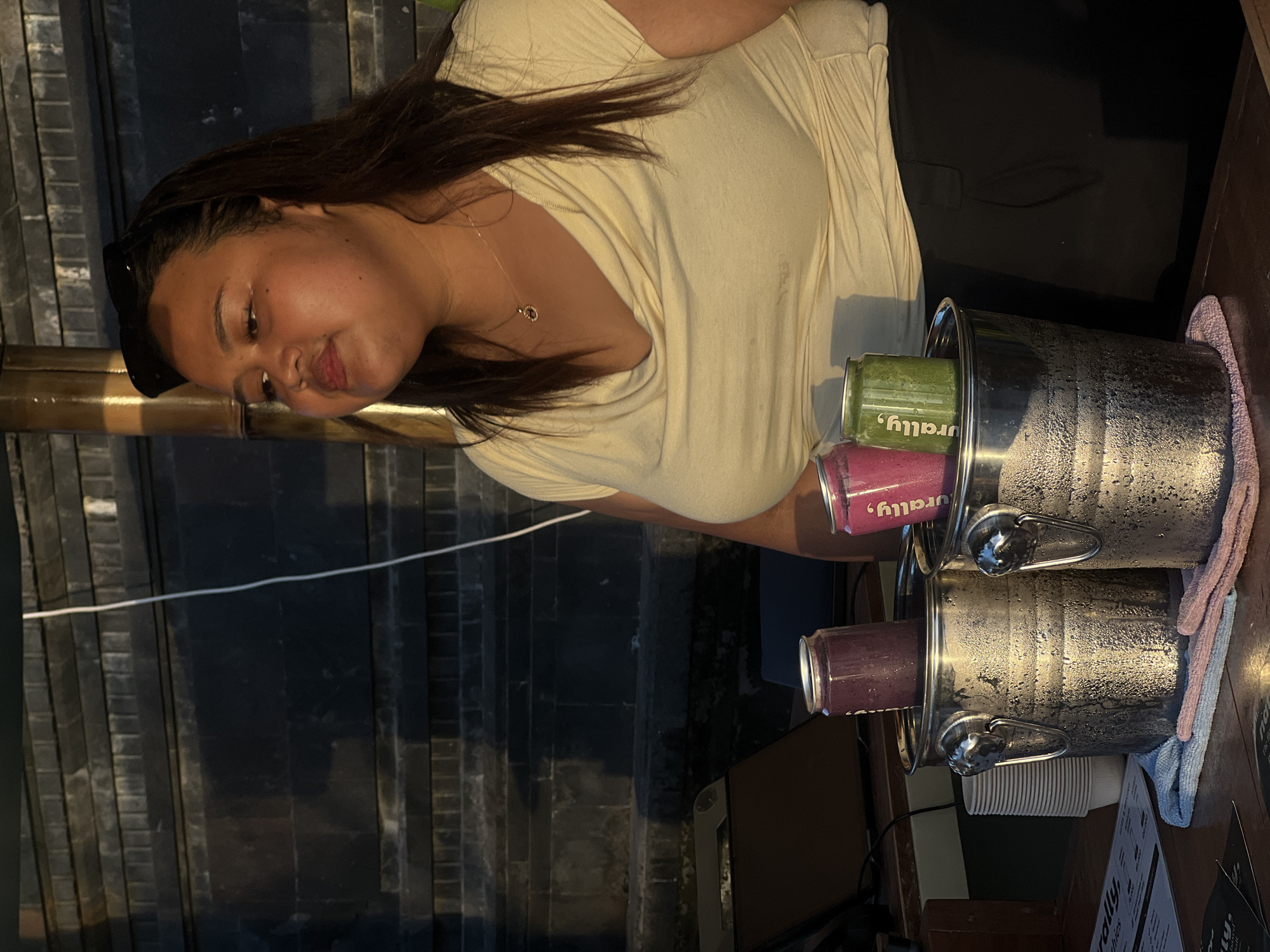 A medium shot captures a woman with dark hair and fair skin from the chest up, wearing a cream-colored cowl-neck t-shirt and black pants. She is standing outdoors, with a stone wall featuring horizontal bands in the background. The woman has sunglasses perched on her head and is looking upwards and to her right. In front of her, two metal ice buckets are filled with condensation and hold two cans of beverages, one purple and one green. A stack of white paper cups is visible to the left of the ice buckets, and a menu lies on the wooden surface in the foreground. The scene suggests a beverage stall in North Kuta, Indonesia, possibly near a beach or outdoor dining area, given the warm lighting and casual attire.