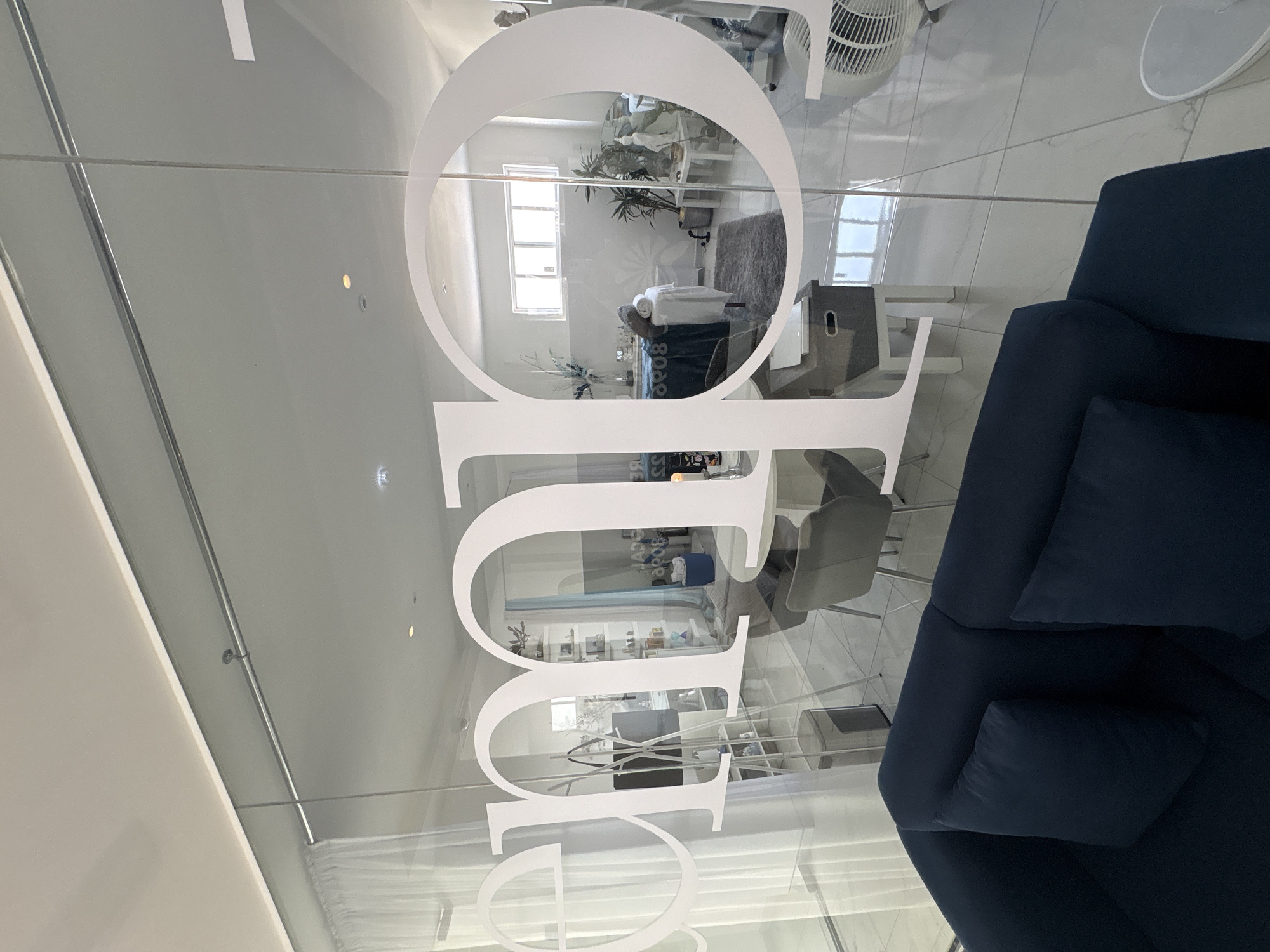 The image provides a forward-facing view into a modern commercial interior, identified as a spa or clinic, located in Puebla, Mexico, observed through a clear glass partition. Large white vinyl letters, including "eme" and "o," are affixed to the glass. In the immediate foreground, the top of a dark blue sofa with two cushions is partially visible.
Beyond the glass, the interior features white tiled flooring and white walls. A dark grey rectangular rug is centrally positioned, on which a treatment bed, covered in a dark teal sheet and topped with neatly folded white towels, rests. To the left, a tall white shelving unit displays various small containers, and a white coat stand holds a dark bag. To the right, a small white table supports a potted plant and a white classical-style bust. A white pedestal fan is located on the floor in the lower right portion of the room. Natural light enters through a horizontal rectangular window in the background, supplemented by recessed ceiling lights. Grey chairs with metal frames are arranged around a small table, and a white console table with a grey storage box is also present. No individuals are depicted within the scene.