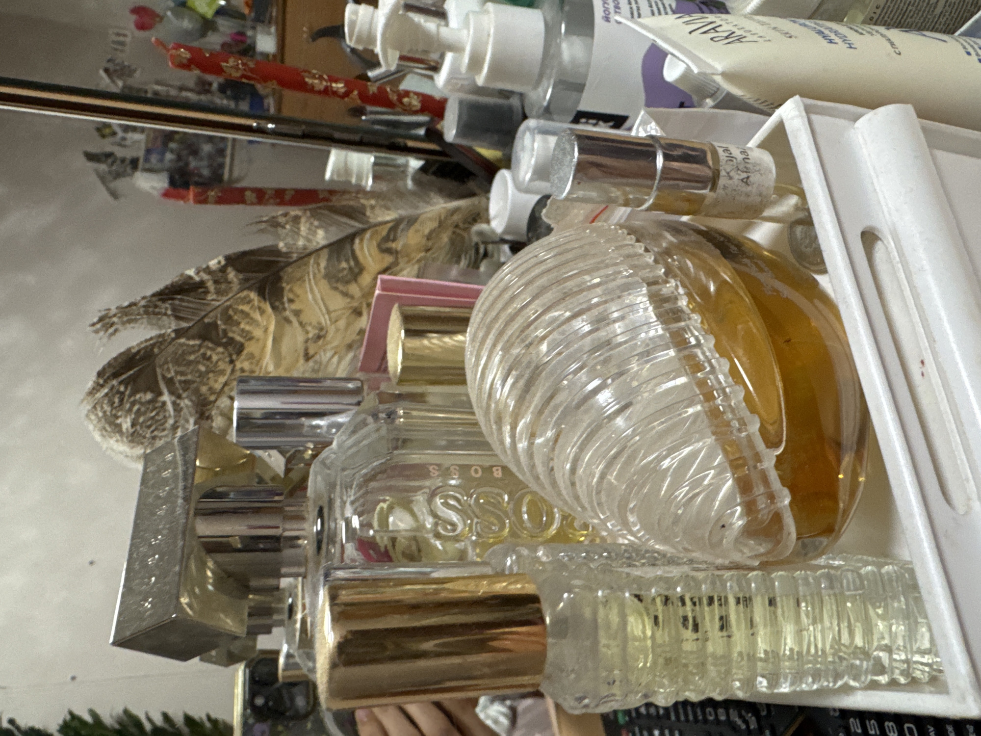 The image shows a collection of personal care items, including various bottles of perfume and lotions, stored in a white plastic container. One of the perfume bottles is distinctly shaped with a clear, spiraled glass design filled with amber liquid. Other bottles feature silver or gold accents and are partially obscured by the main perfume bottle. A patterned feather is visible in the background. The items are densely packed within the container. There is no evidence that points to the location being in Izhevsk, Russia.