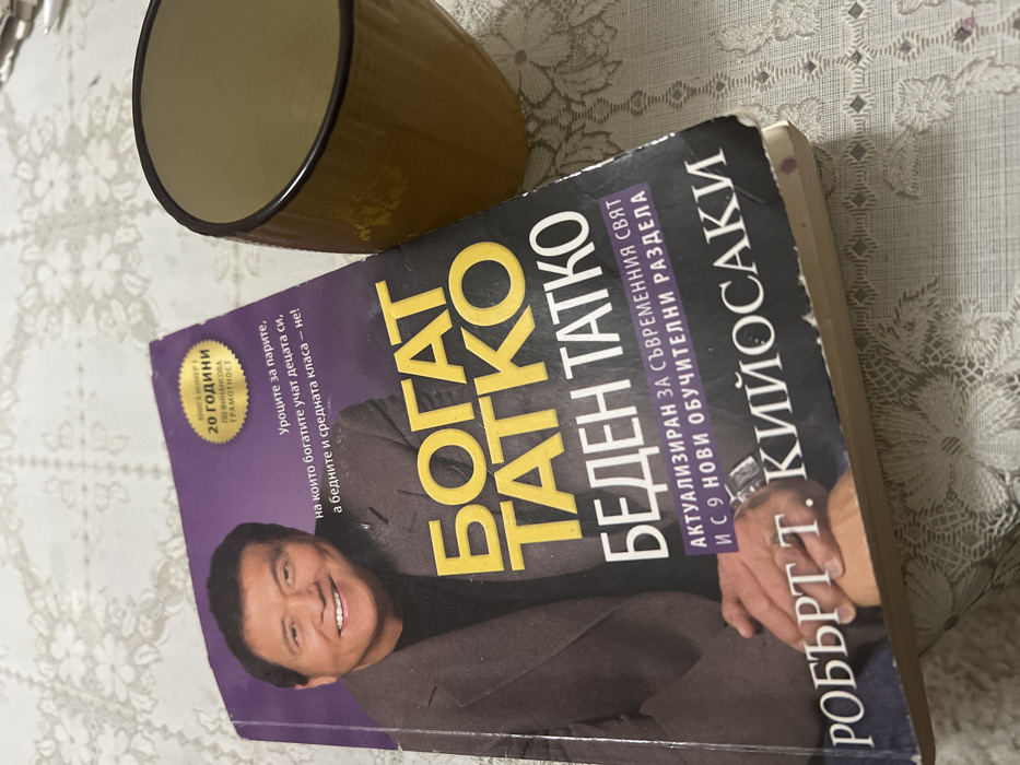 A Bulgarian edition of the book "Rich Dad Poor Dad" by Robert T. Kiyosaki, titled "БОГАТ ТАТКО БЕДЕН ТАТКО," is centrally positioned on a patterned white tablecloth. The book cover features a smiling Robert Kiyosaki in a dark suit, with yellow and white Cyrillic text. A circular yellow seal on the upper right of the cover reads "20 ГОДИНИ ПО ФИНАНСОВА ГРАМОТНОСТ" (20 YEARS OF FINANCIAL LITERACY). The book exhibits minor wear along its edges and spine. To the right of the book, a mustard-yellow ceramic mug with a dark brown rim stands, appearing empty. The tablecloth features a repeating floral or star-like white lace pattern. The scene is illuminated by overhead light. The photograph was taken in Sedlartsi, Bulgaria.