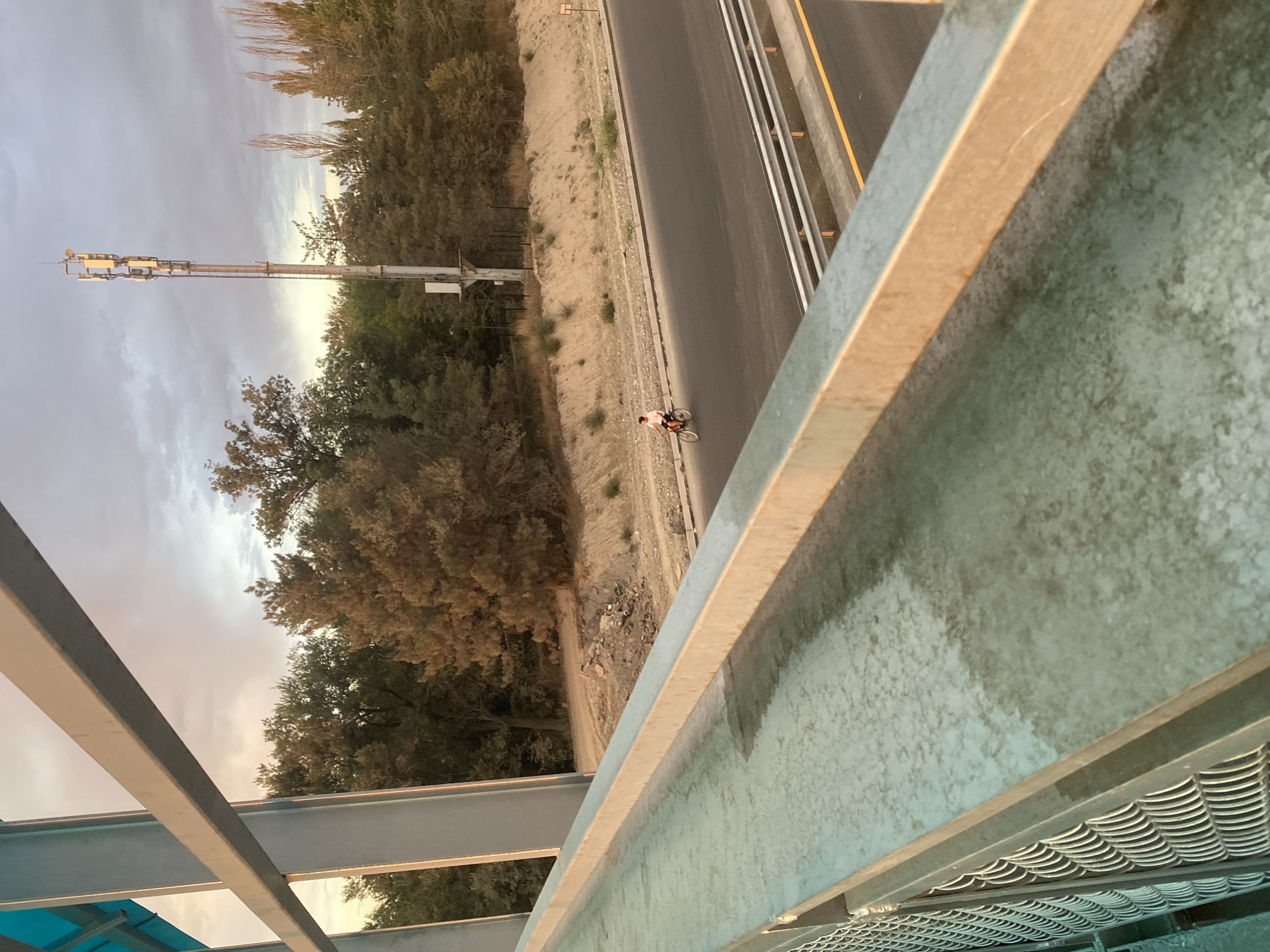 A person is seen riding a bicycle on a multi-lane road. The road appears to be a highway or a major thoroughfare, with guardrails and clear lane markings. The surrounding environment includes a steep, sandy embankment on one side, dotted with dry vegetation and trees. A tall telecommunications tower stands prominently against a cloudy sky. The scene is framed by the perspective of someone looking through a metal railing, suggesting the vantage point is from an overpass or a bridge. The lighting indicates it's either early morning or late afternoon, with warm hues reflecting off the metal structures. The overall setting suggests a semi-urban or fringe-of-city location, possibly within the greater Bishkek area, given the presence of infrastructure like the telecommunications tower and the highway.